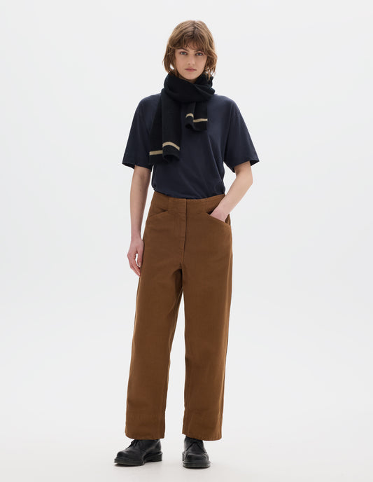 Full body image, front view of model wearing a dark short sleeve t shirt, brown cotton trousers, with front pockets and deep hems. Styled with a black wool scarf with beige edge stripe and black leather shoes.