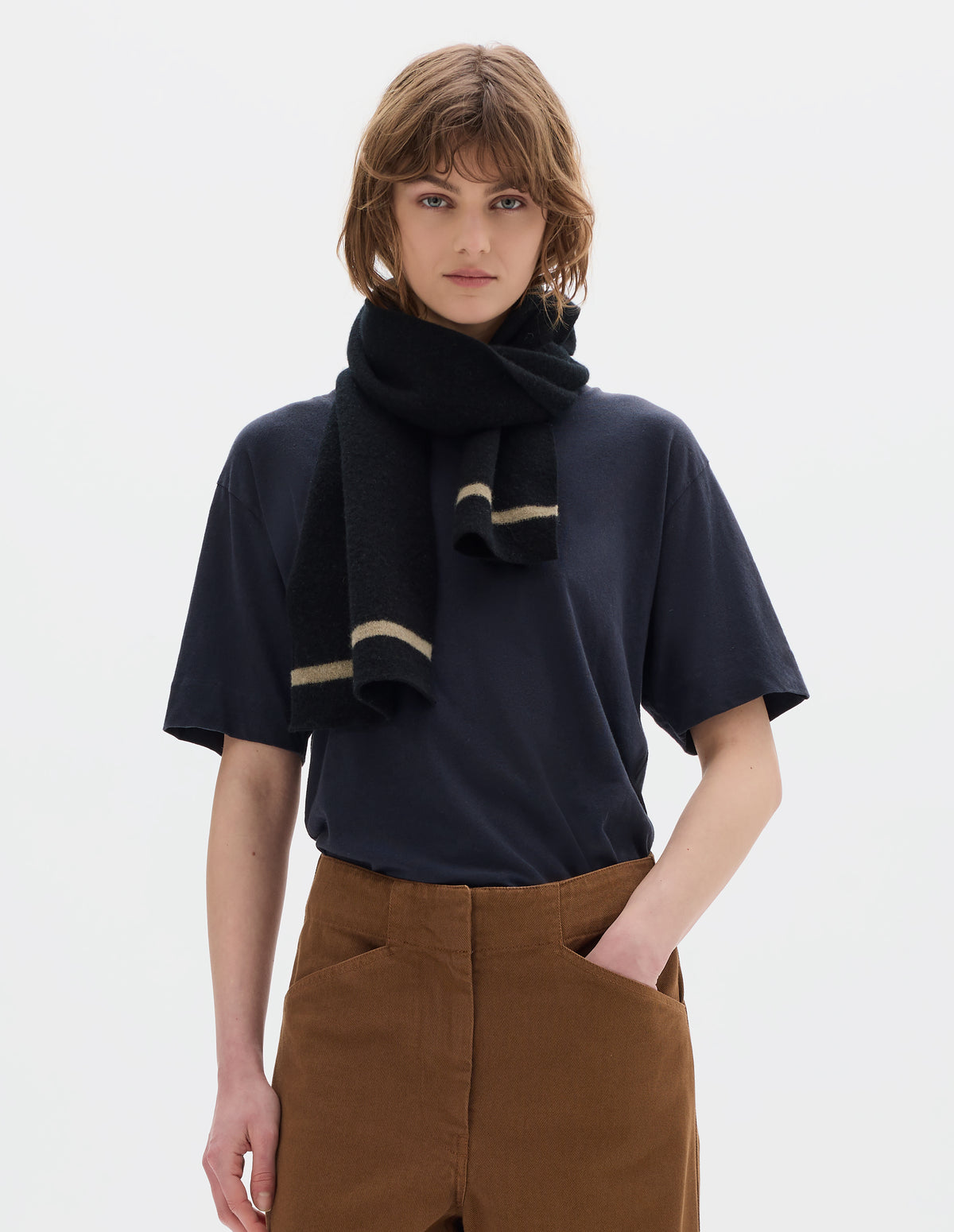 Cropped body image, front view of model wearing a black lambswool scarf with a beige stripe, styled with a short sleeve navy t shirt and orangey brown cotton trousers.