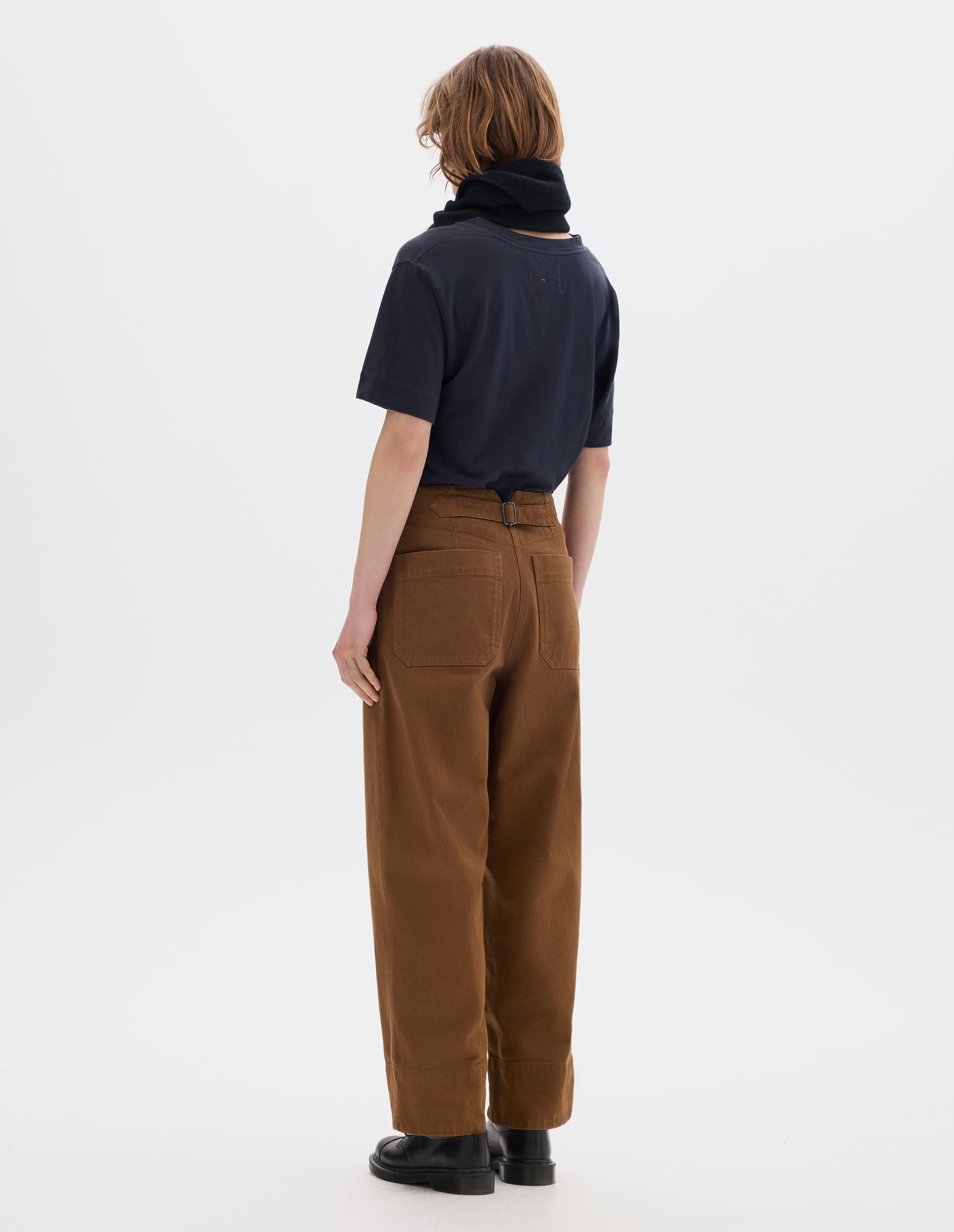 Full body image, back view of model wearing dark short sleeve t shirt, brown cotton trousers, with back patch pockets, cinch waist adjuster and deep hems. Styled with a black wool scarf and black leather shoes.
