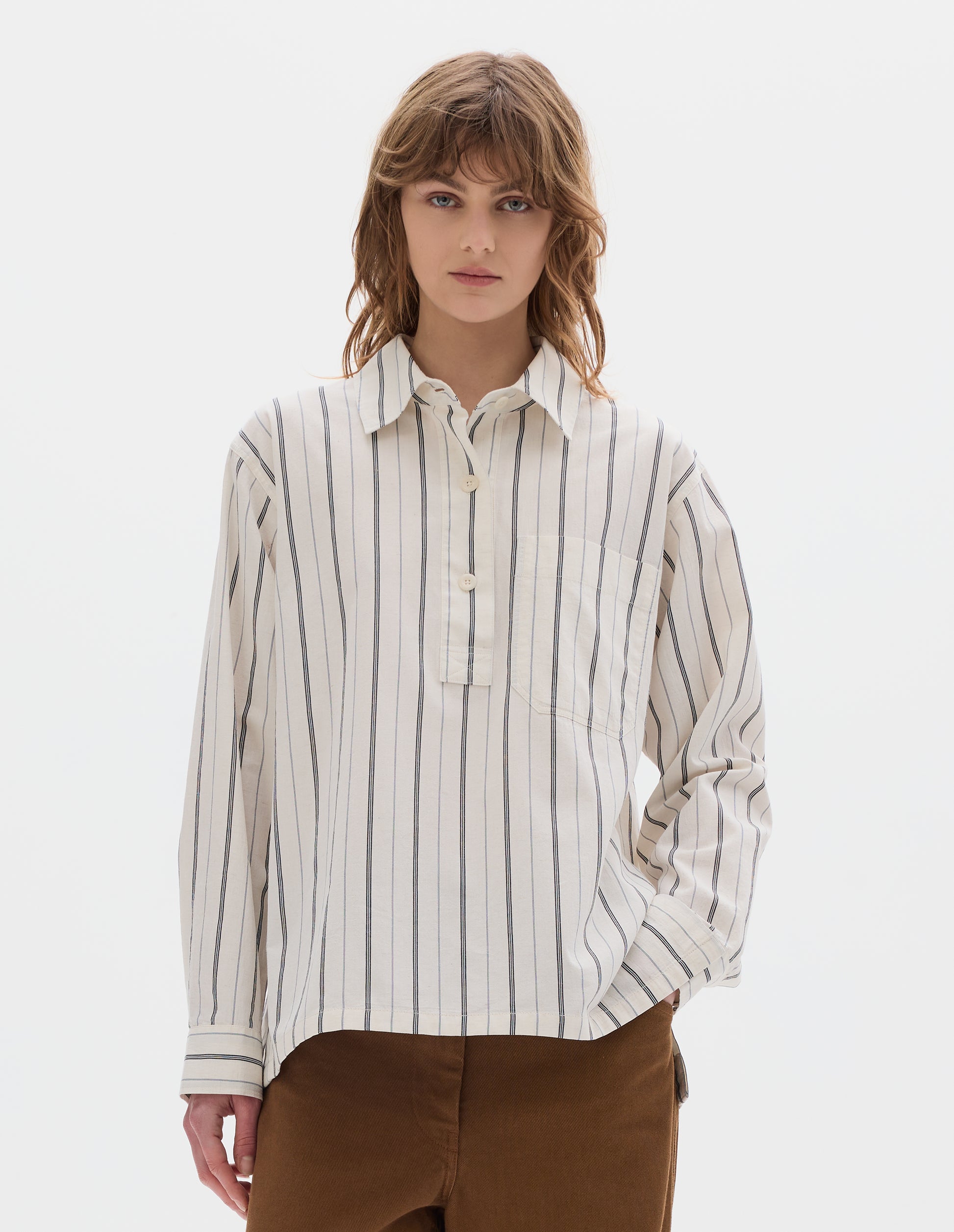 Cropped body image, front view of model wearing an off white and dark blue stripe cotton shirt with light buttons,  a half placket, chest patch pocket. Styled with brown tan trousers.
