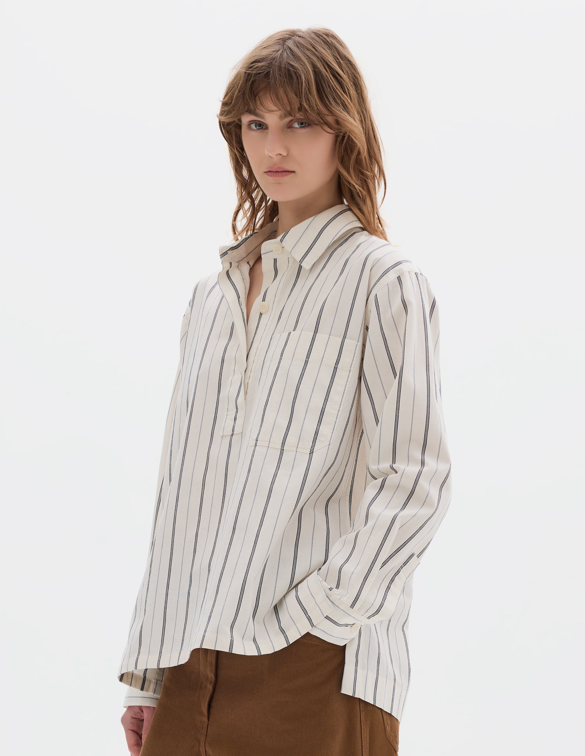 Cropped body image, side view of model wearing an off white and dark blue stripe cotton shirt with light buttons, a half placket, chest patch pocket, with deep side splits and a stepped hem. Styled with brown tan trousers.