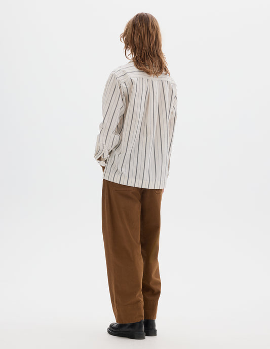 Full body image, back view of model wearing an off white and dark blue stripe cotton. Box pleat on back yoke. Styled with brown tan trousers and black leather shoes.