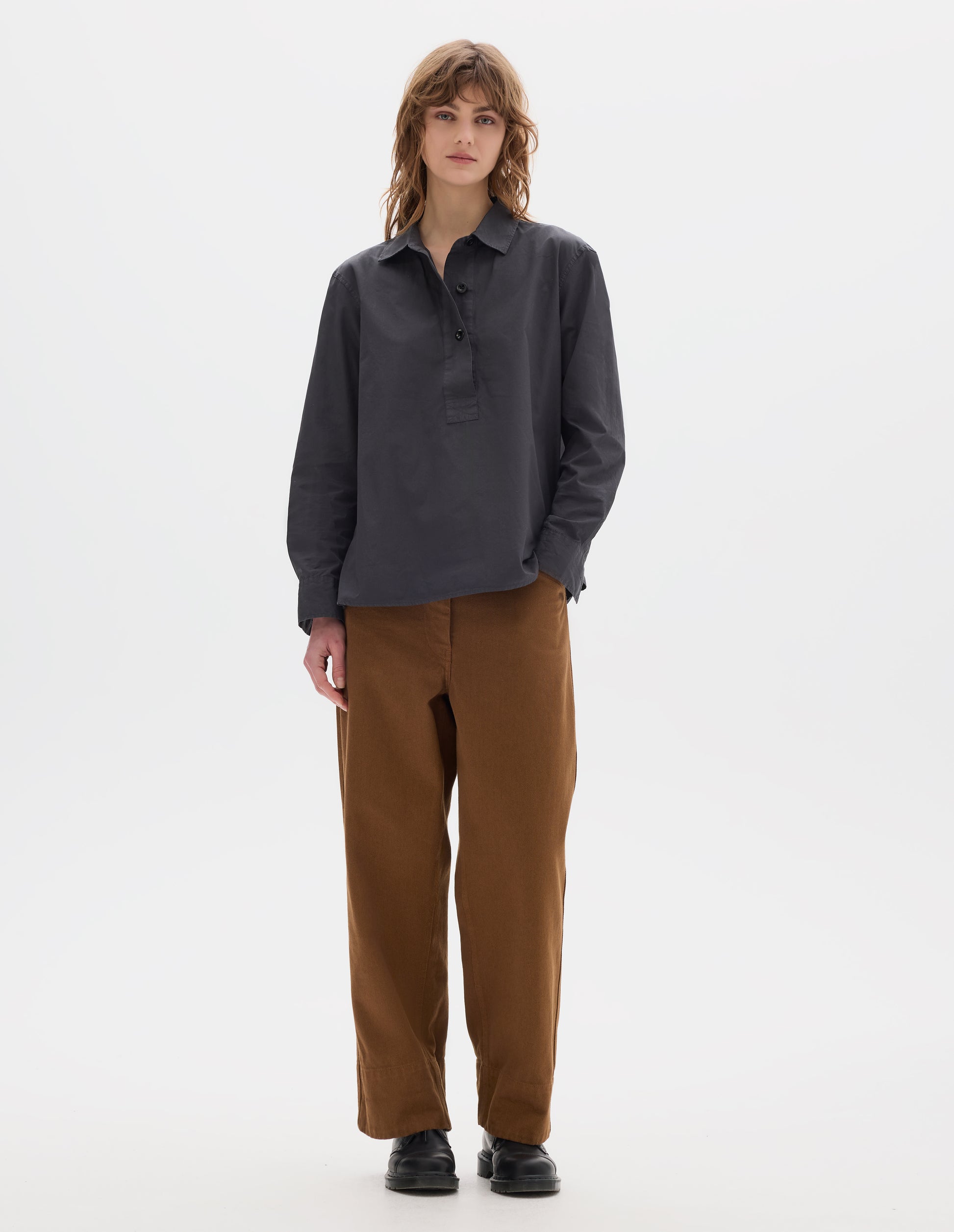 Full body image, front view of model wearing a dark grey shirt with an A line cut, constructed with a raised half placket. Styled with brown tan trousers and black leather shoes.