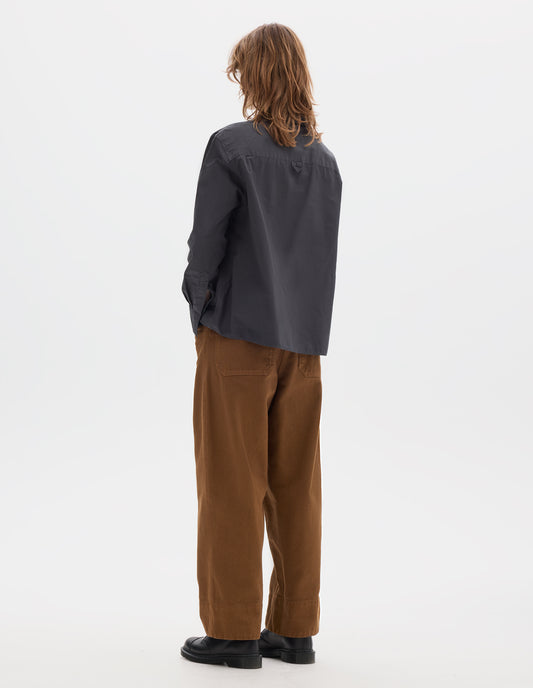Full body image, back view of model wearing dark grey shirt with an A line cut with and a hanger loop detail on the back yoke. Styled with brown tan trousers and black leather shoes.