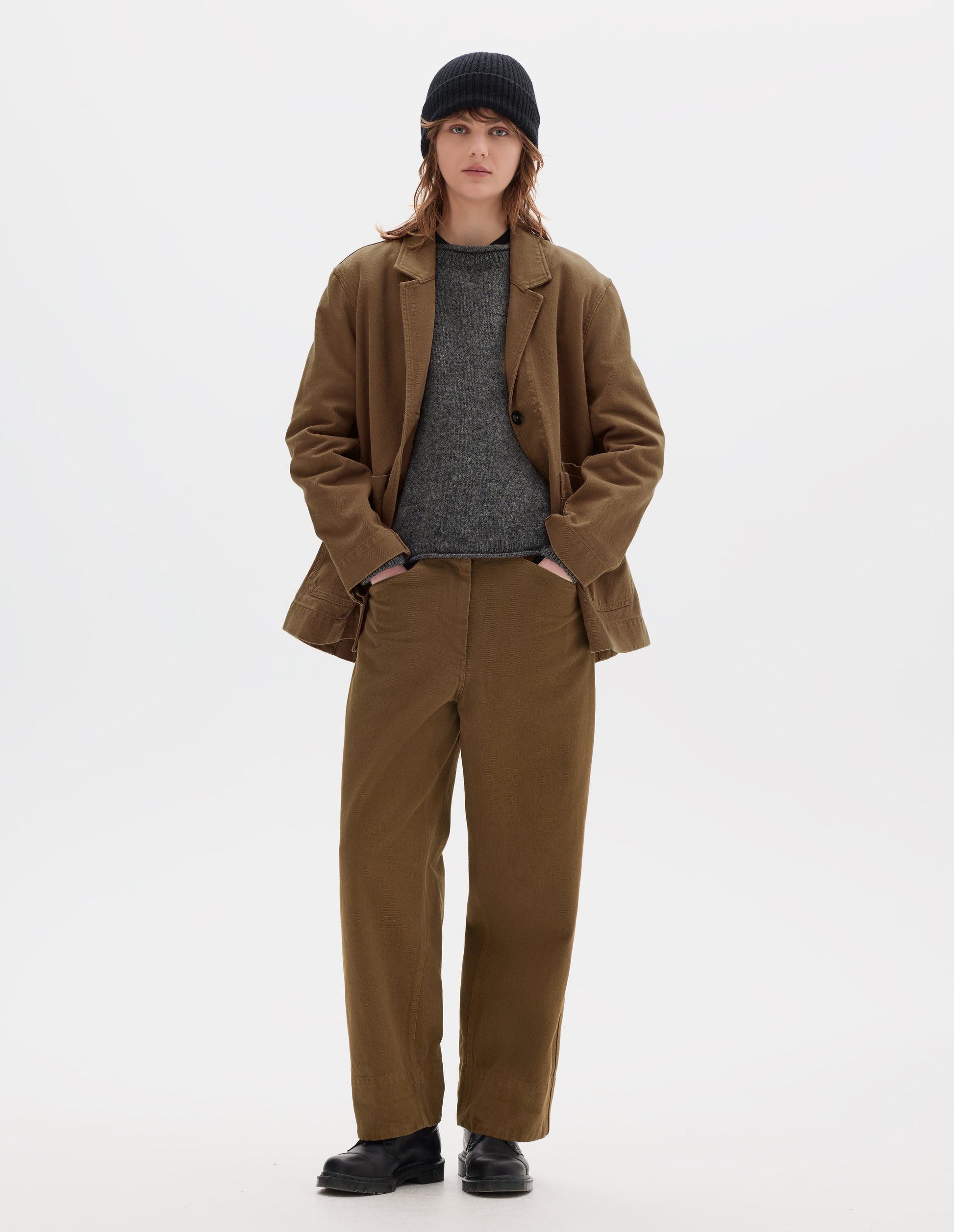 Full body image, front view of model wearing a brown cotton blazer, worn open over a grey wool jumper. Styled with a black wool beanie, matching brown flat front straight leg cotton trousers and black leather shoes.