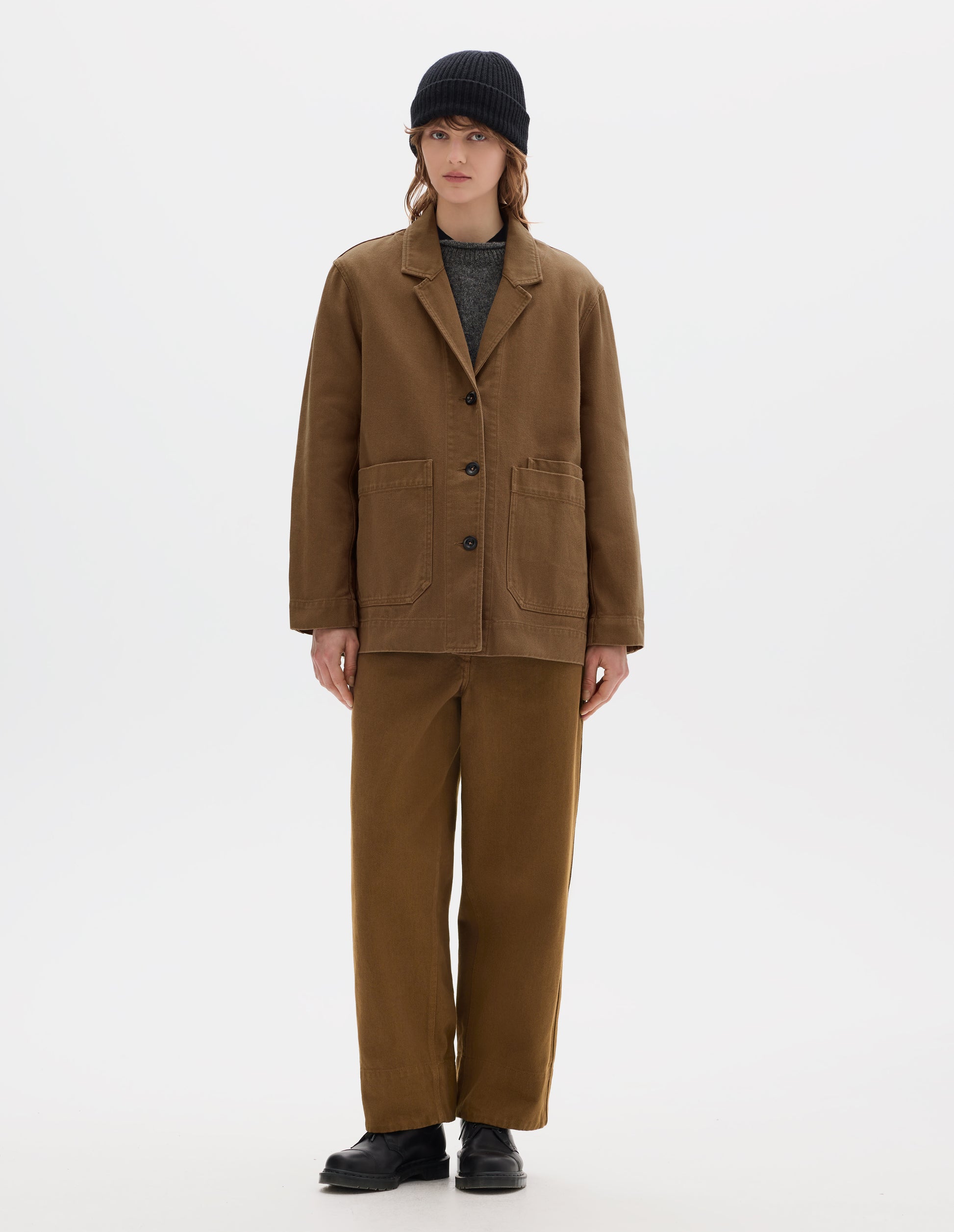 Full body image, front view of model wearing a brown cotton blazer, with large front pockets and three dark brown buttons, worn over a grey wool jumper. Styled with a black wool beanie, matching brown straight leg cotton trousers and black leather shoes.