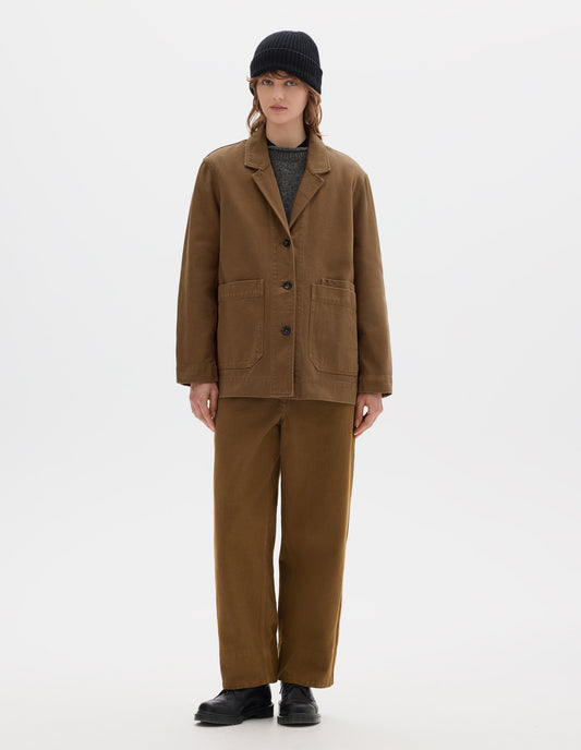 Full body image, front view of model wearing a brown cotton blazer, with large front pockets and three dark brown buttons, worn over a grey wool jumper. Styled with a black wool beanie, matching brown straight leg cotton trousers and black leather shoes.