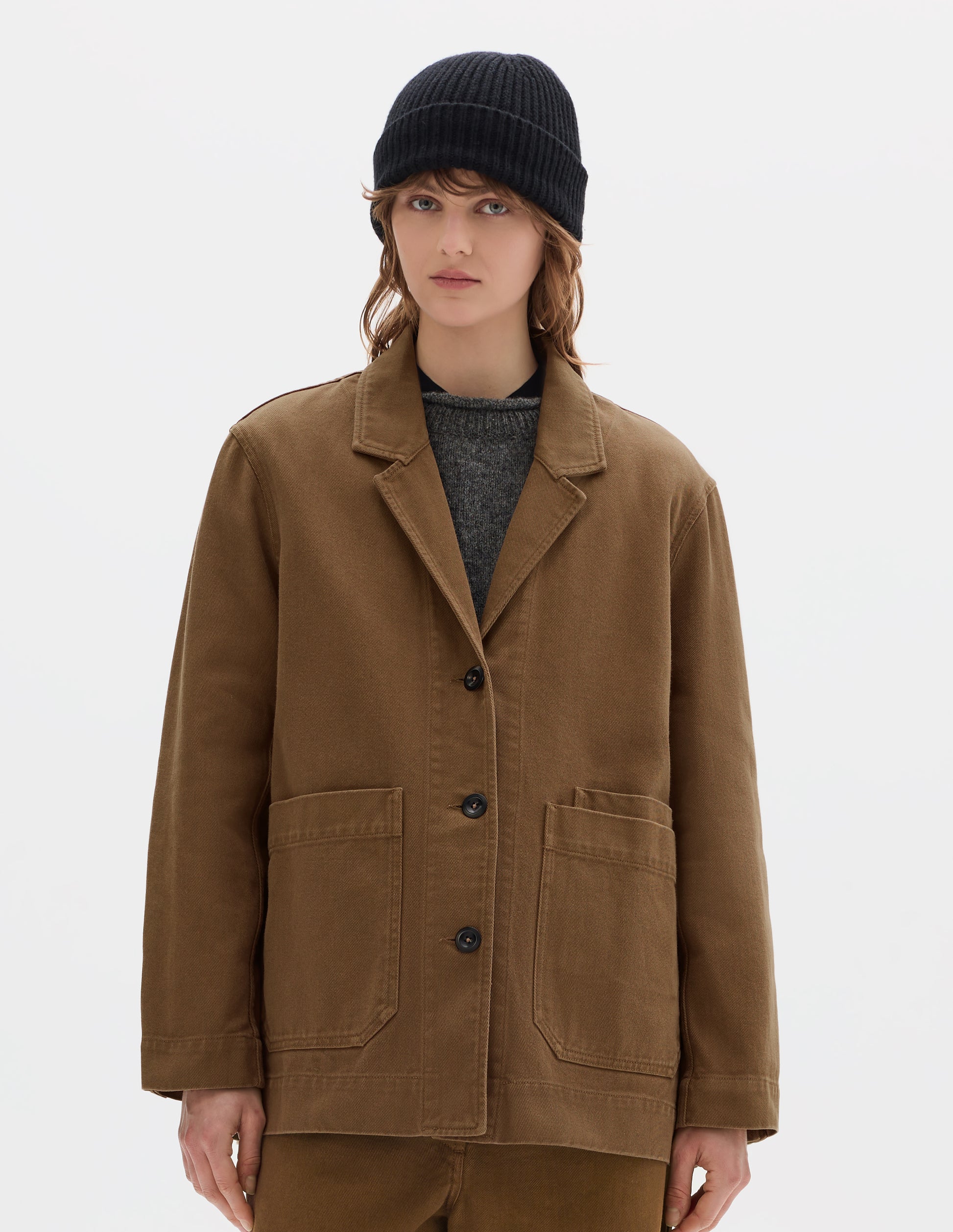 Cropped body image, front view of model wearing a brown cotton blazer, with large front pockets and three dark brown buttons, worn over a grey wool jumper. Styled with a black wool beanie and matching brown cotton trousers.