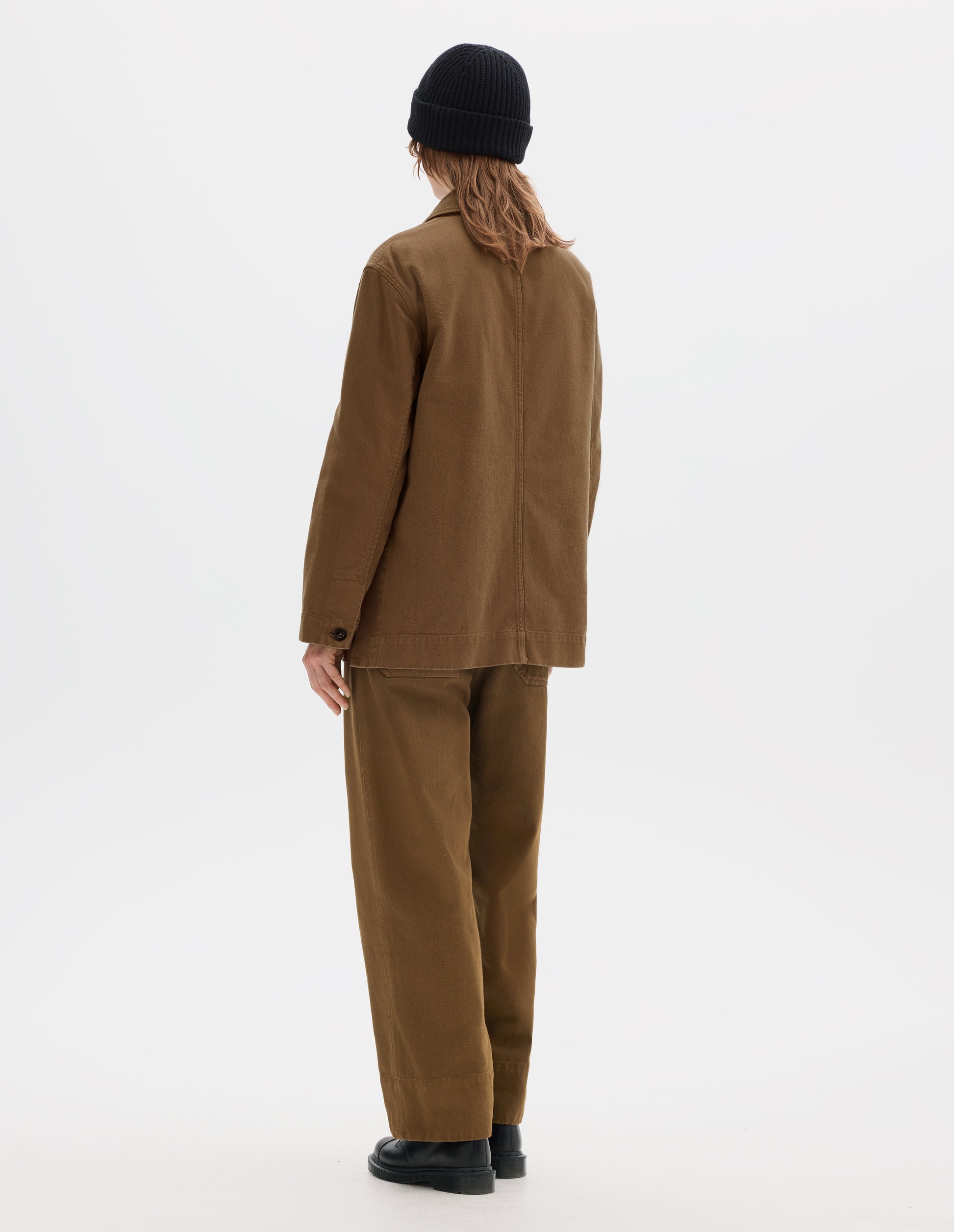 Full body image, back view of model wearing a a brown cotton blazer which features grown-on cuffs with a wide placket opening, centre back seam detailing. Styled with a black wool beanie, matching brown straight leg cotton trousers and black leather shoes.