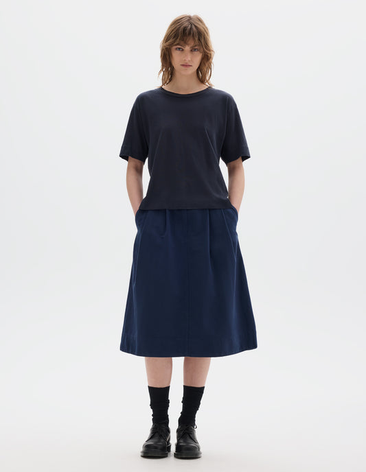 Full body image, front view of model wearing navy blue short sleeve cotton t-shirt with contrast rib on neck. Styled with indigo blue pleated skirt, black socks and leather shoes.