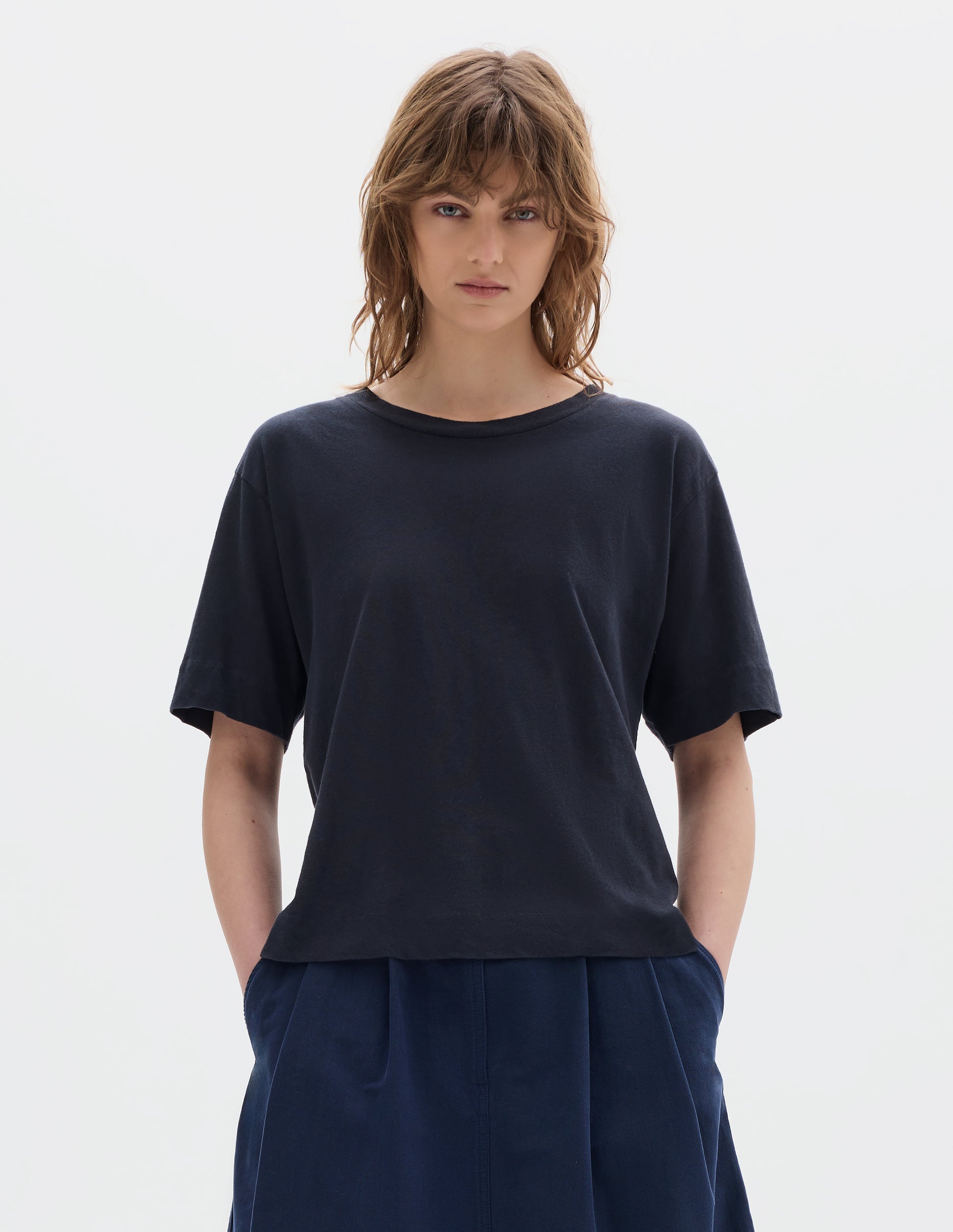 Cropped body image, front view of model wearing navy blue short sleeve cotton t-shirt with contrast rib on neck. Styled with indigo blue pleated skirt.