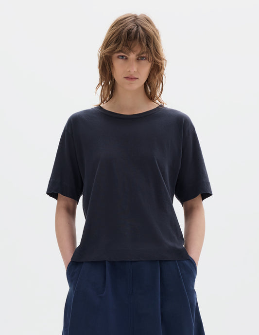 Cropped body image, front view of model wearing navy blue short sleeve cotton t-shirt with contrast rib on neck. Styled with indigo blue pleated skirt.