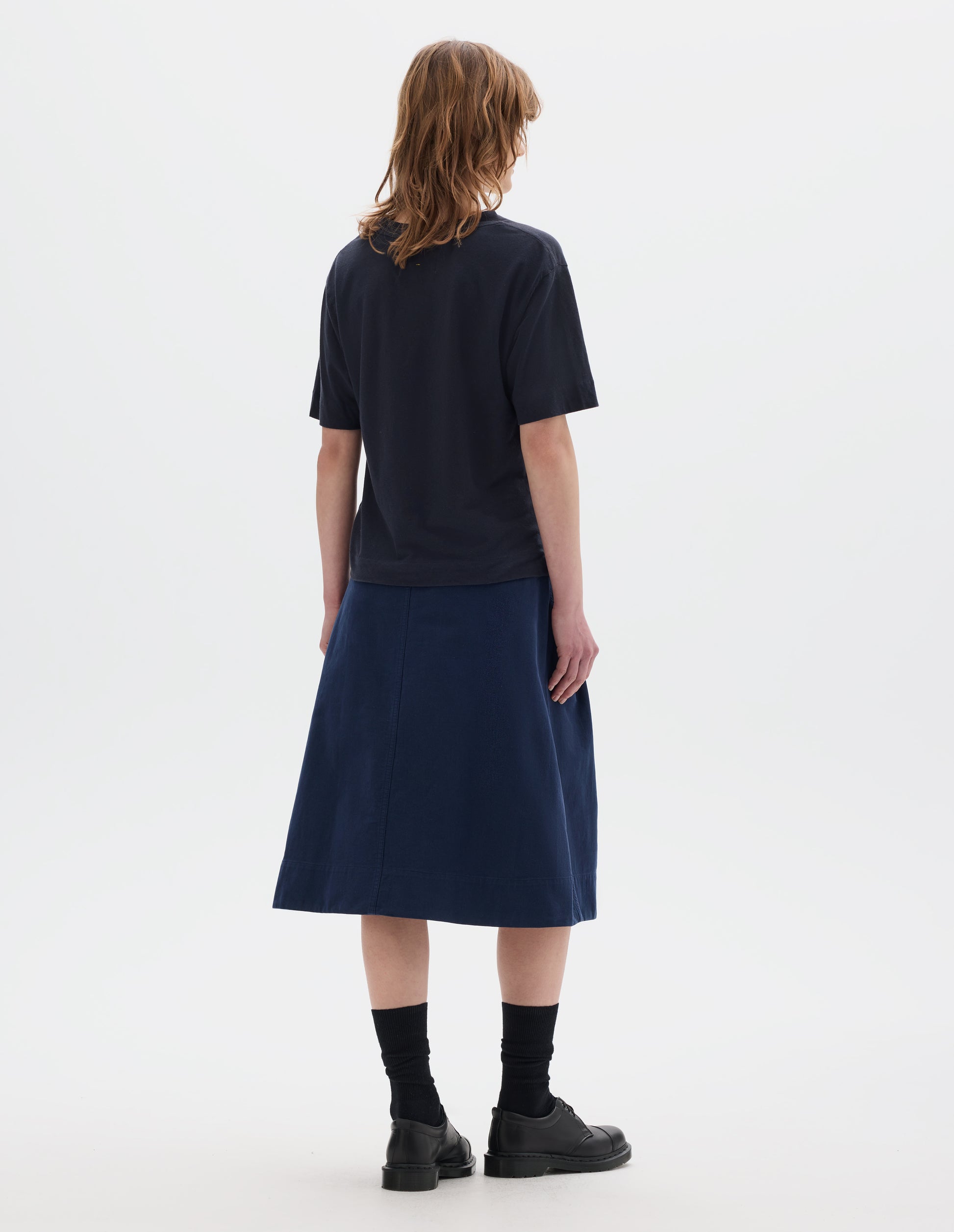 Full body image, back view of model wearing navy blue short sleeve cotton t-shirt with contrast rib on neck and stitch detailing on back shoulder. Styled with indigo blue skirt, black socks and leather shoes.