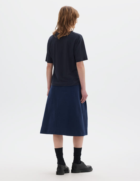 Full body image, back view of model wearing navy blue short sleeve cotton t-shirt with contrast rib on neck and stitch detailing on back shoulder. Styled with indigo blue skirt, black socks and leather shoes.