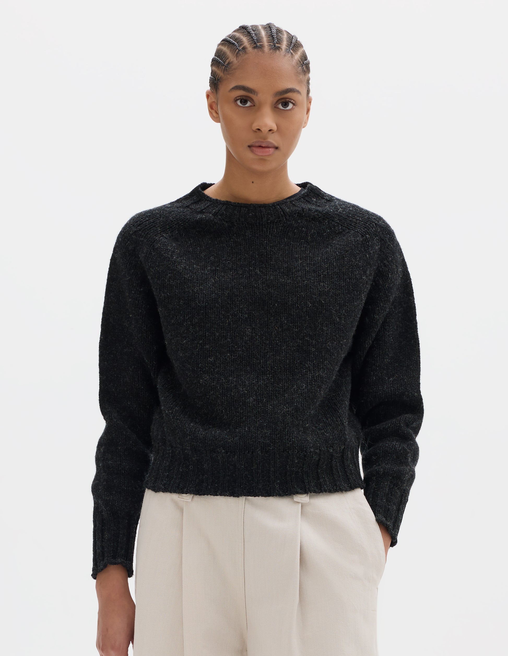 Cropped body image, front view of model wearing dark grey wool jumper with saddle sleeve and contrast rib on neckline, cuffs and hem. Styled with off white pleated trousers.
