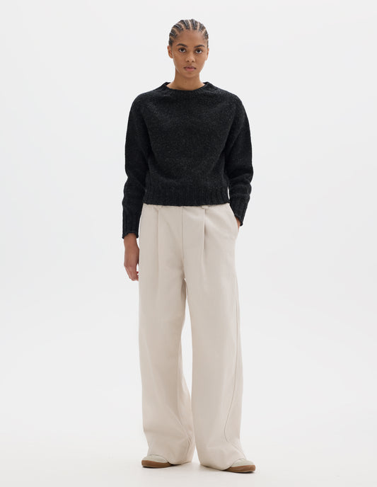 Full body image, front view of model wearing dark grey wool jumper with saddle sleeve and contrast rib on neckline, cuffs and hem. Styled with off white pleated trousers and white trainers with gum sole.