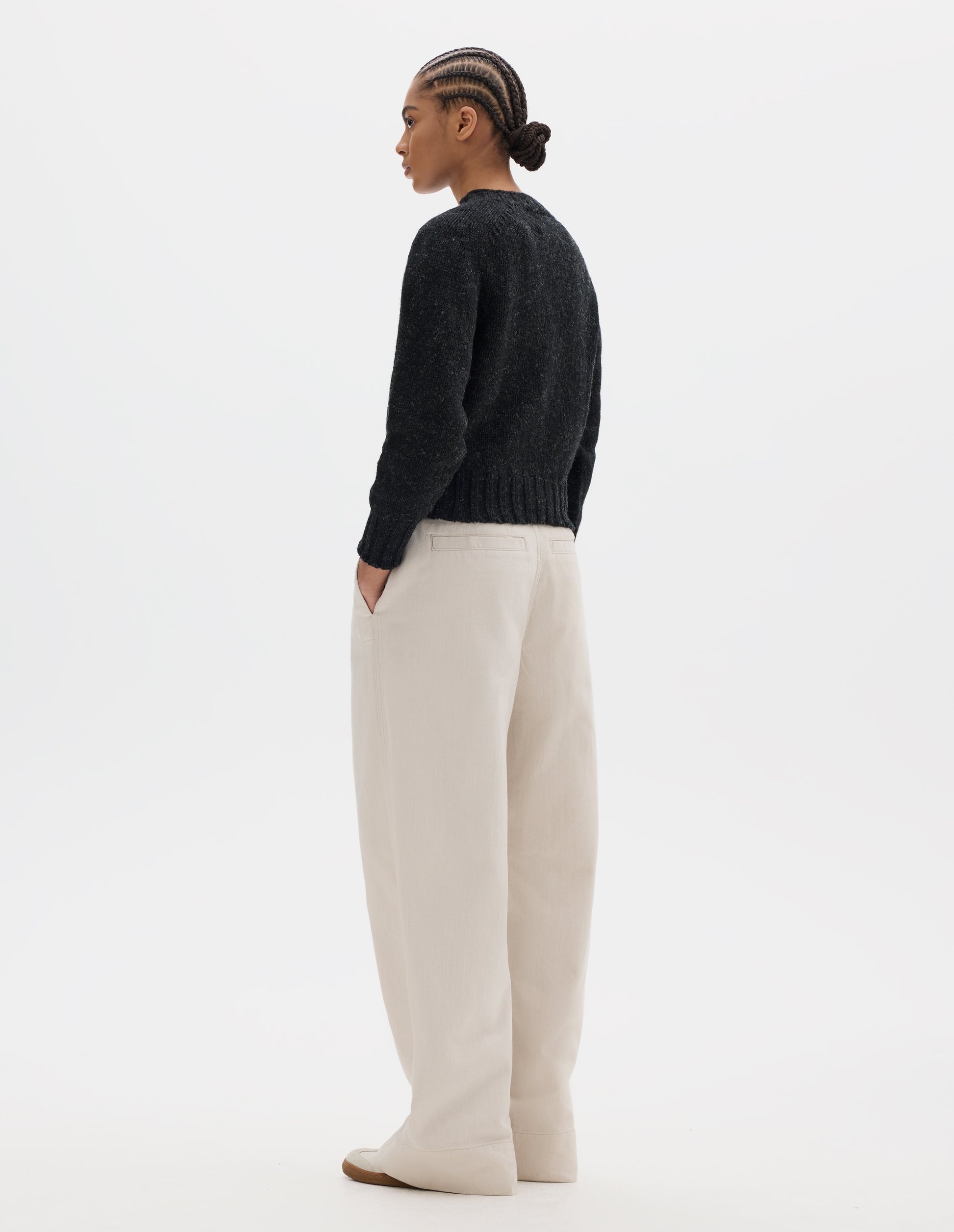 Full body image, back view of model wearing dark grey wool jumper with saddle sleeve and contrast rib on neckline, cuffs and hem. Styled with off white pleated trousers and white trainers with gum sole.