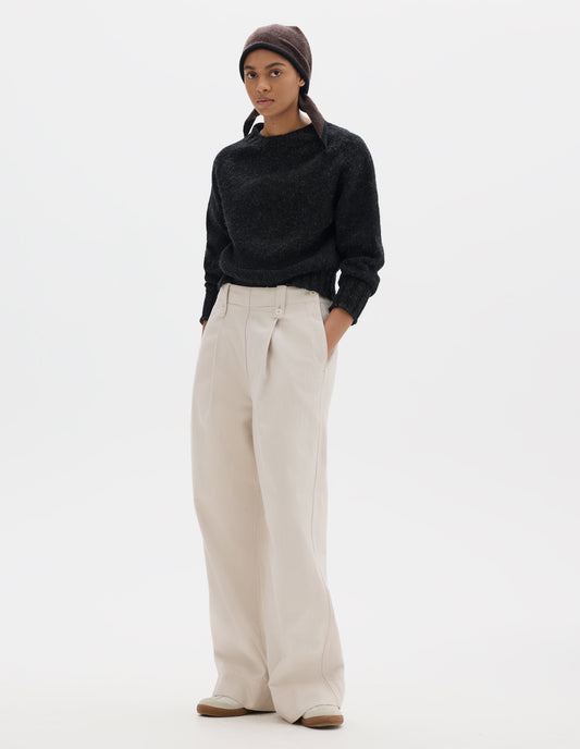 Full body image, front view of model wearing off white trousers with with grown-on waistband, button-fastening belt loops, a side opening with integrated side seam pockets, and two front pleats. Styled with dark grey jumped tucked in, brown knitted scarf worn on head and white trainers with gum sole.