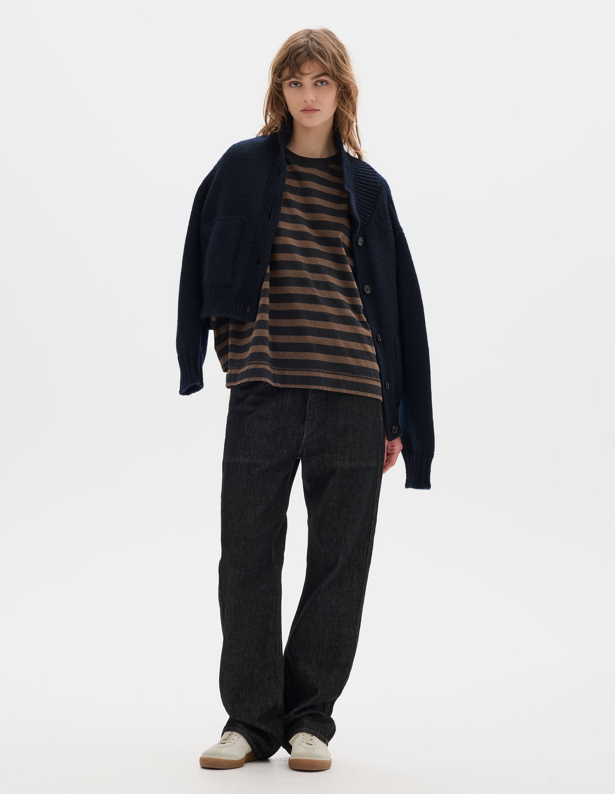 Full body image, front view of model wearing a dark navy blue boxy fit collared cardigan, with laid-on side entry pockets, and rib detailing on collar, placket, hem and cuffs. Worn open, styled with a striped brown and black jersey top, black denim trousers and off white suede trainers.