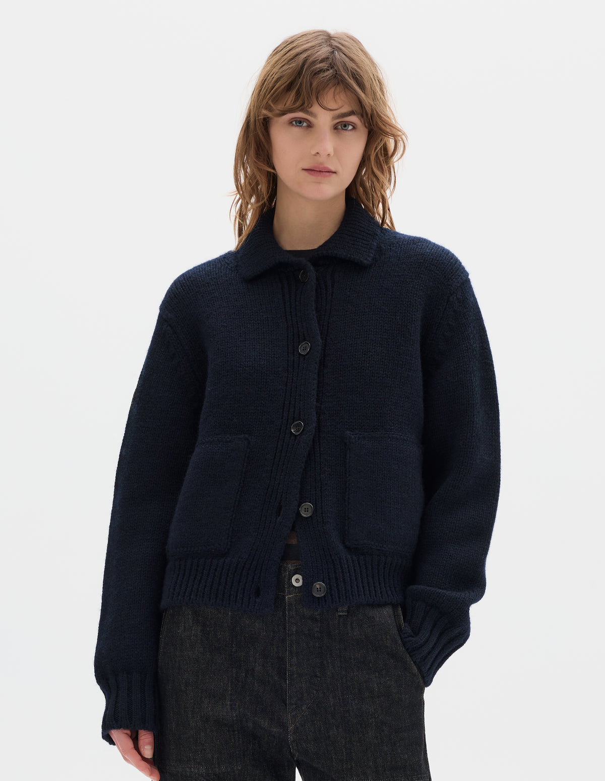 Cropped body image, front view of model wearing a dark navy blue boxy fit collared cardigan, with laid-on side entry pockets, and rib detailing on collar, placket, hem and cuffs. Styled with black denim trousers and worn over a striped jersey.
