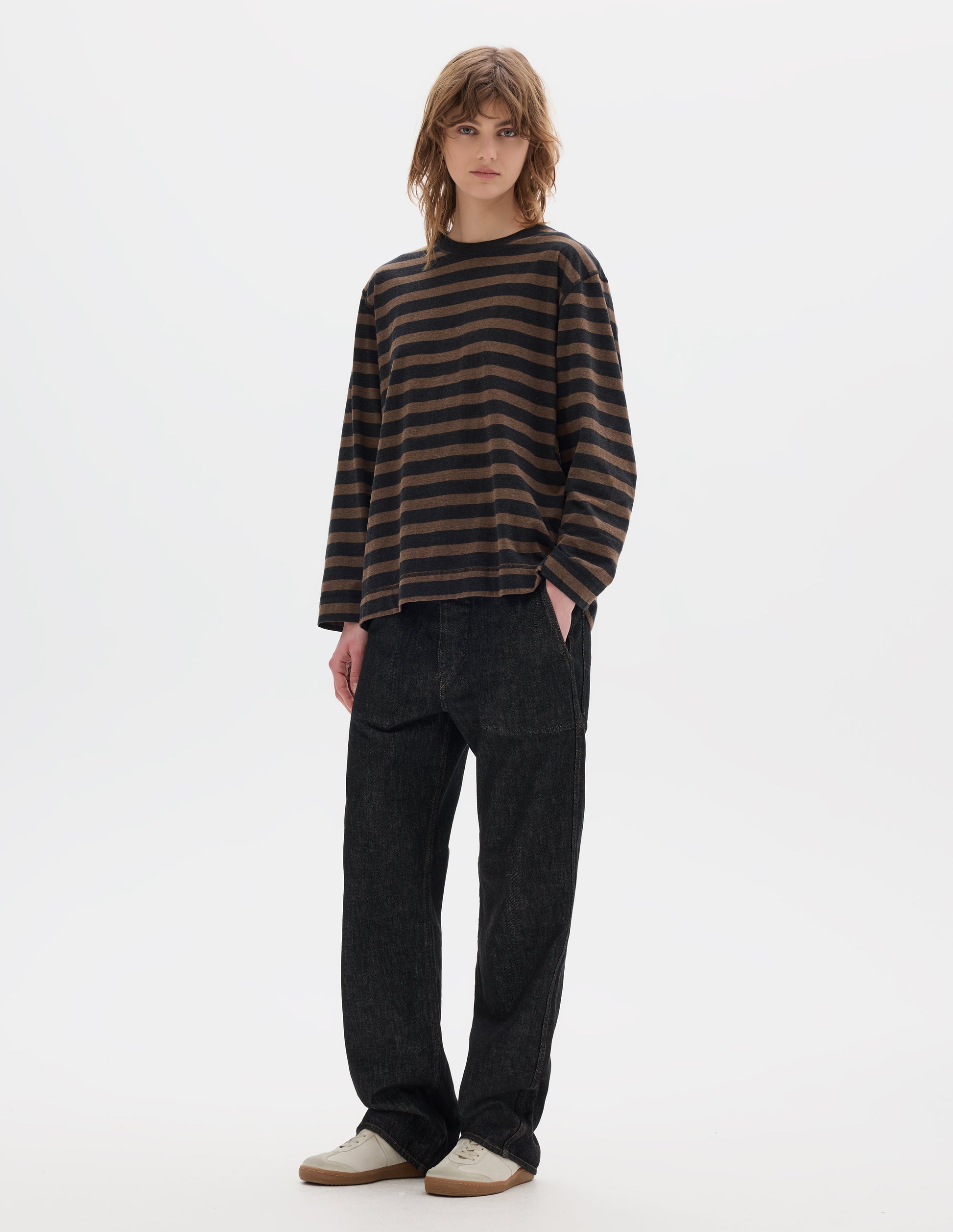 Full body image, front view of model wearing off black and brown stripe cotton top, with contrasting rib neck and flatlock stitch detailing on the seams. Styled with black jeans and white shoes.