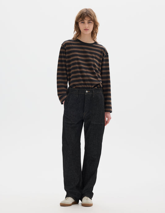 Full body image, front view of model wearing black denim twill jeans, with metal button fly, stitch-through waistband with belt loops, and stitch through pockets. Styled with black and brown striped matelot t shirt and white suede trainers. 
