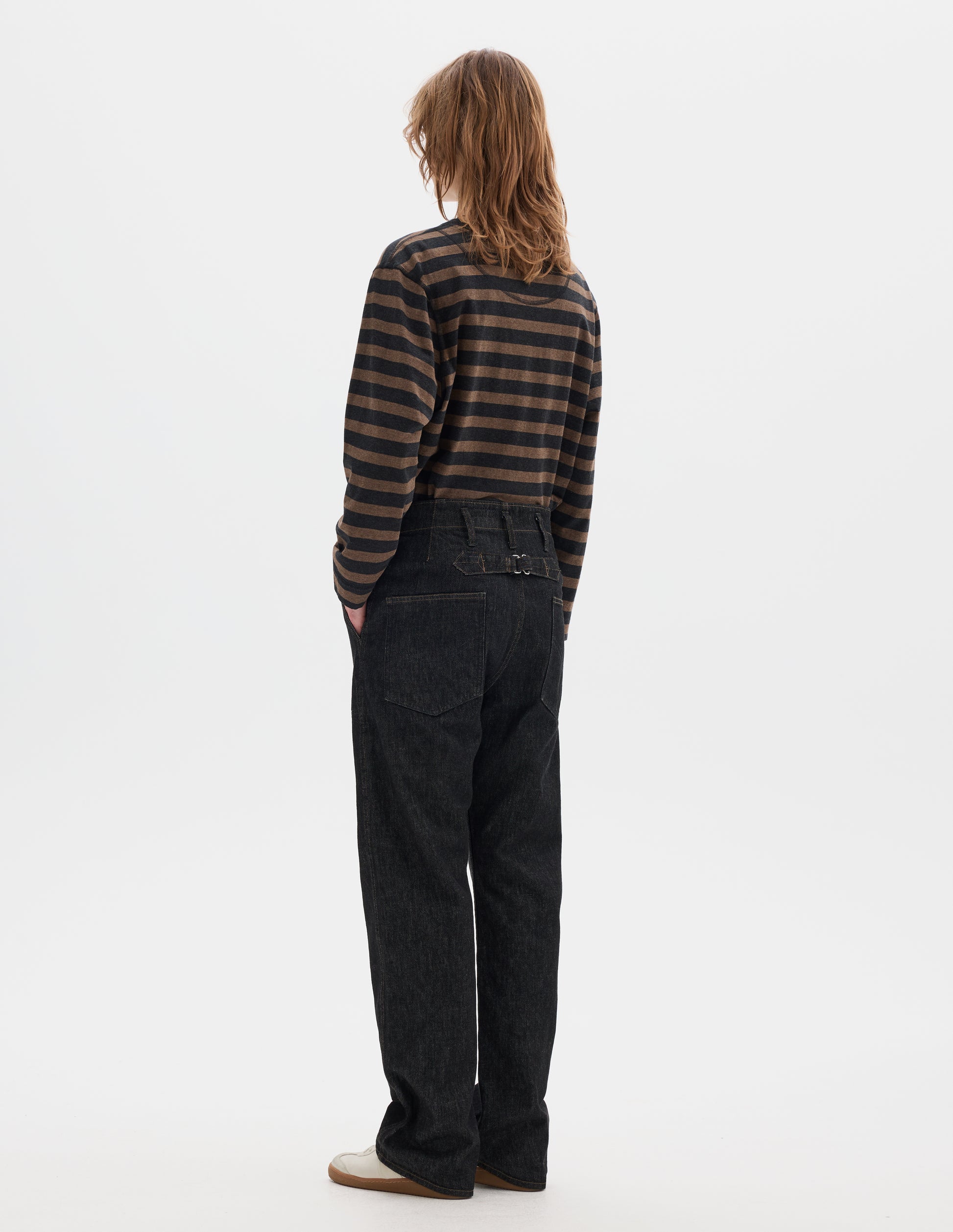 Full body image, back view of model wearing black denim twill jeans, with stitch-through waistband with belt loops, cinch back detail with metal buckle and patch back pockets. Styled with black and brown striped matelot t shirt and white suede trainers. 