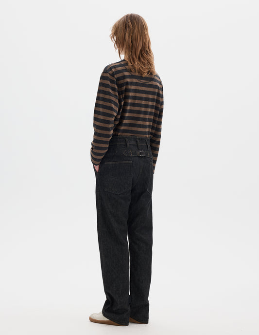 Full body image, back view of model wearing black denim twill jeans, with stitch-through waistband with belt loops, cinch back detail with metal buckle and patch back pockets. Styled with black and brown striped matelot t shirt and white suede trainers. 