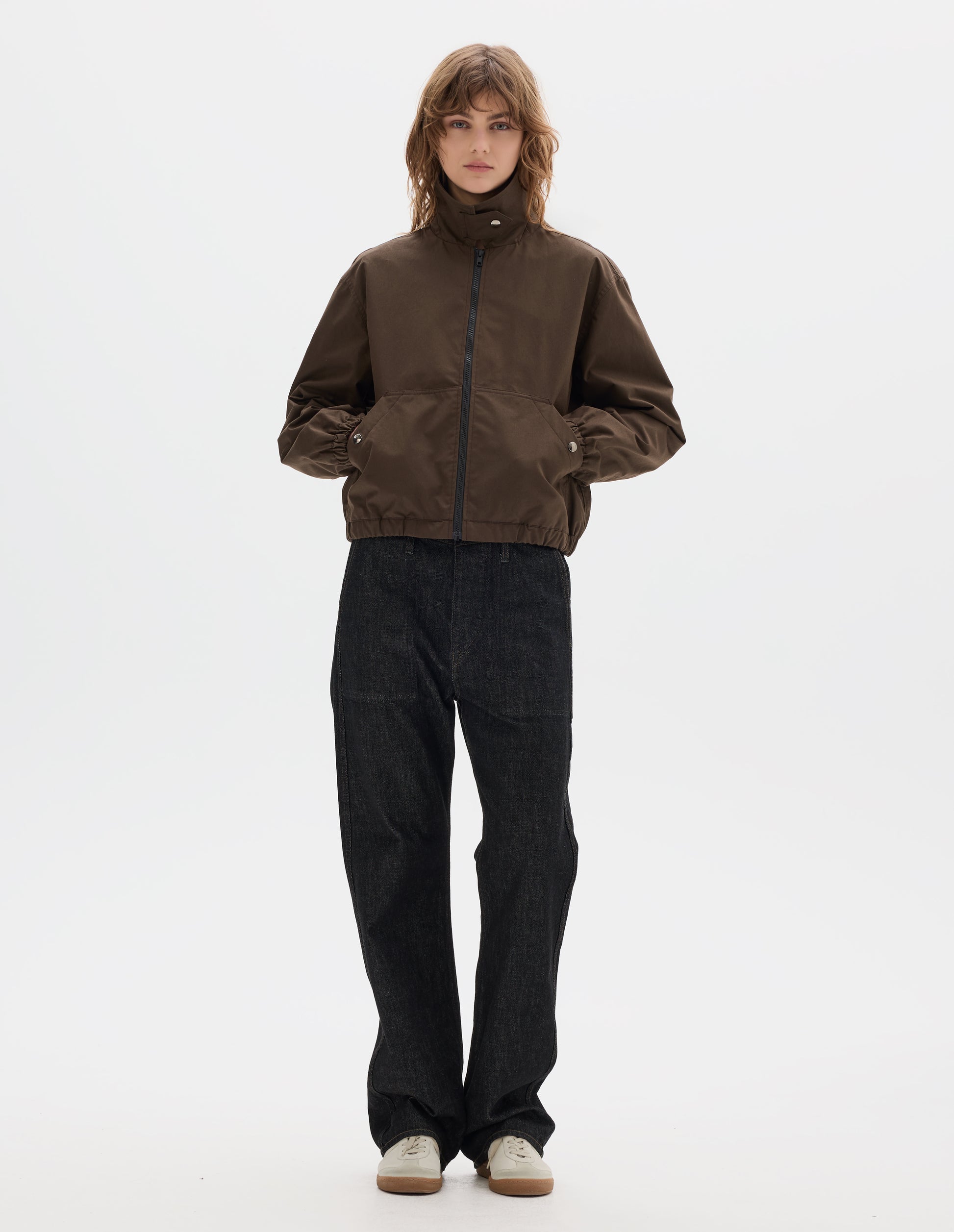 Full body image, front view of model wearing a fastened brown zip-through front jacket, cropped in length, a collar with a throat tab and press stud fastening done up, two slanted front pockets fastened with press studs, hands in pockets, and an elasticated half-hem and cuffs. Styled with black denim and pale beige trainers.