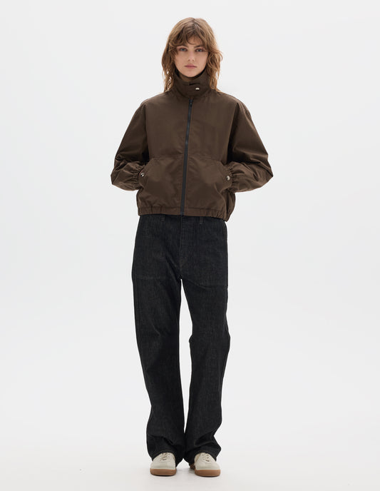 Full body image, front view of model wearing a fastened brown zip-through front jacket, cropped in length, a collar with a throat tab and press stud fastening done up, two slanted front pockets fastened with press studs, hands in pockets, and an elasticated half-hem and cuffs. Styled with black denim and pale beige trainers.