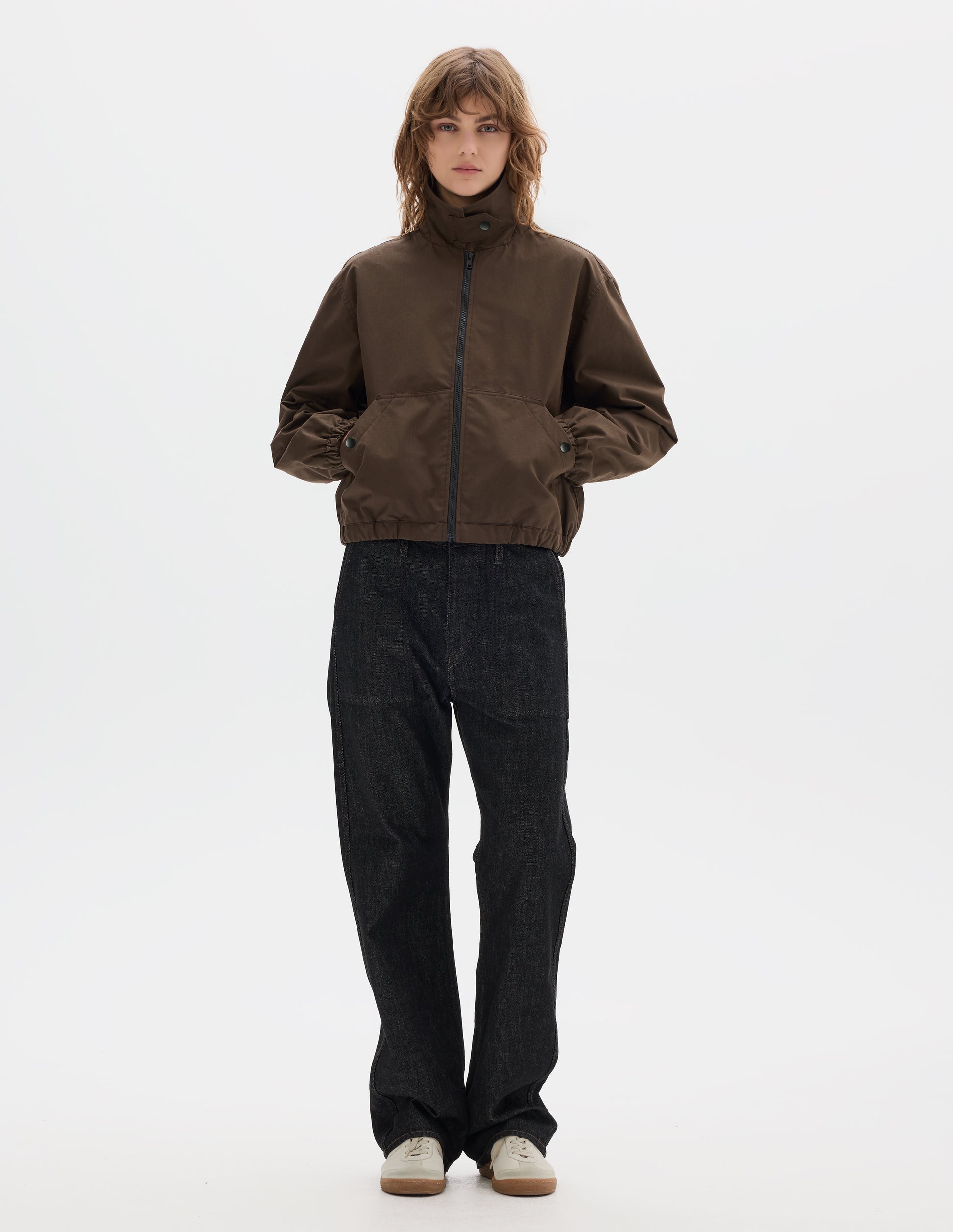 Full body image, front view of model wearing a fastened brown zip-through front jacket, cropped in length, a collar with a throat tab and press stud fastening done up, two slanted front pockets fastened with press studs, hands in pockets, and an elasticated half-hem and cuffs. Styled with black denim and pale beige trainers.