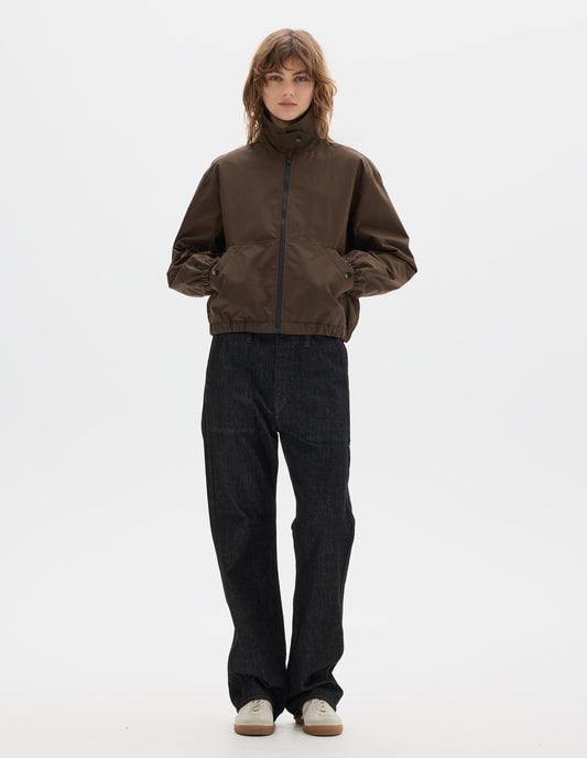 Full body image, front view of model wearing a fastened brown zip-through front jacket, cropped in length, a collar with a throat tab and press stud fastening done up, two slanted front pockets fastened with press studs, hands in pockets, and an elasticated half-hem and cuffs. Styled with black denim and pale beige trainers.