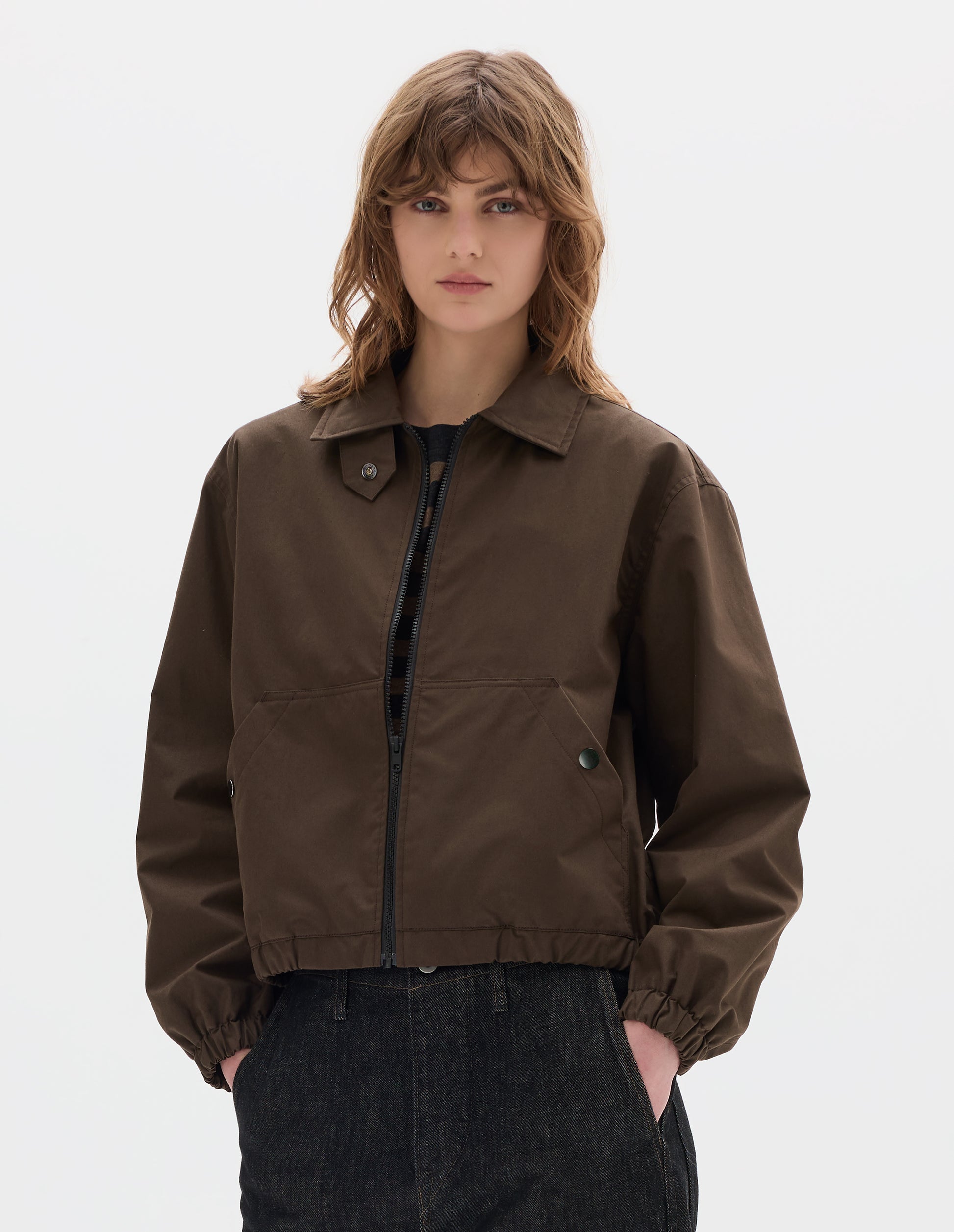 Cropped body image, front view of model wearing an open brown zip-through front jacket, cropped in length, a collar with a throat tab and press stud fastening, two slanted front pockets fastened with press studs, and an elasticated half-hem and cuffs. Styled with black denim.