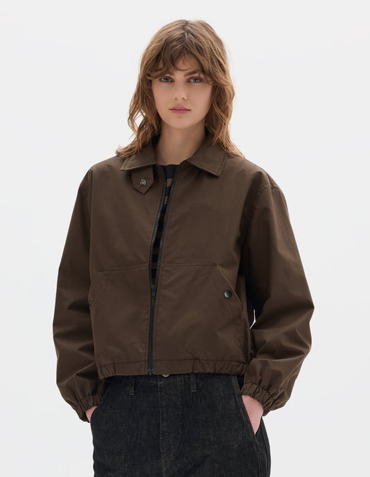 Cropped body image, front view of model wearing an open brown zip-through front jacket, cropped in length, a collar with a throat tab and press stud fastening, two slanted front pockets fastened with press studs, and an elasticated half-hem and cuffs. Styled with black denim.