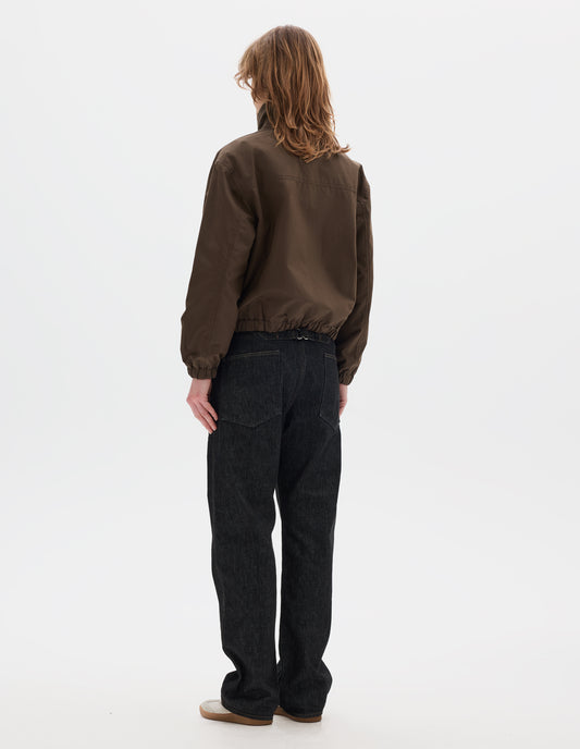 Full body image, back view of model wearing a  brown jacket, cropped in length, a collar with a throat tab and press stud fastening done up,   and an elasticated hem and cuffs. Styled with black denim and pale beige trainers.