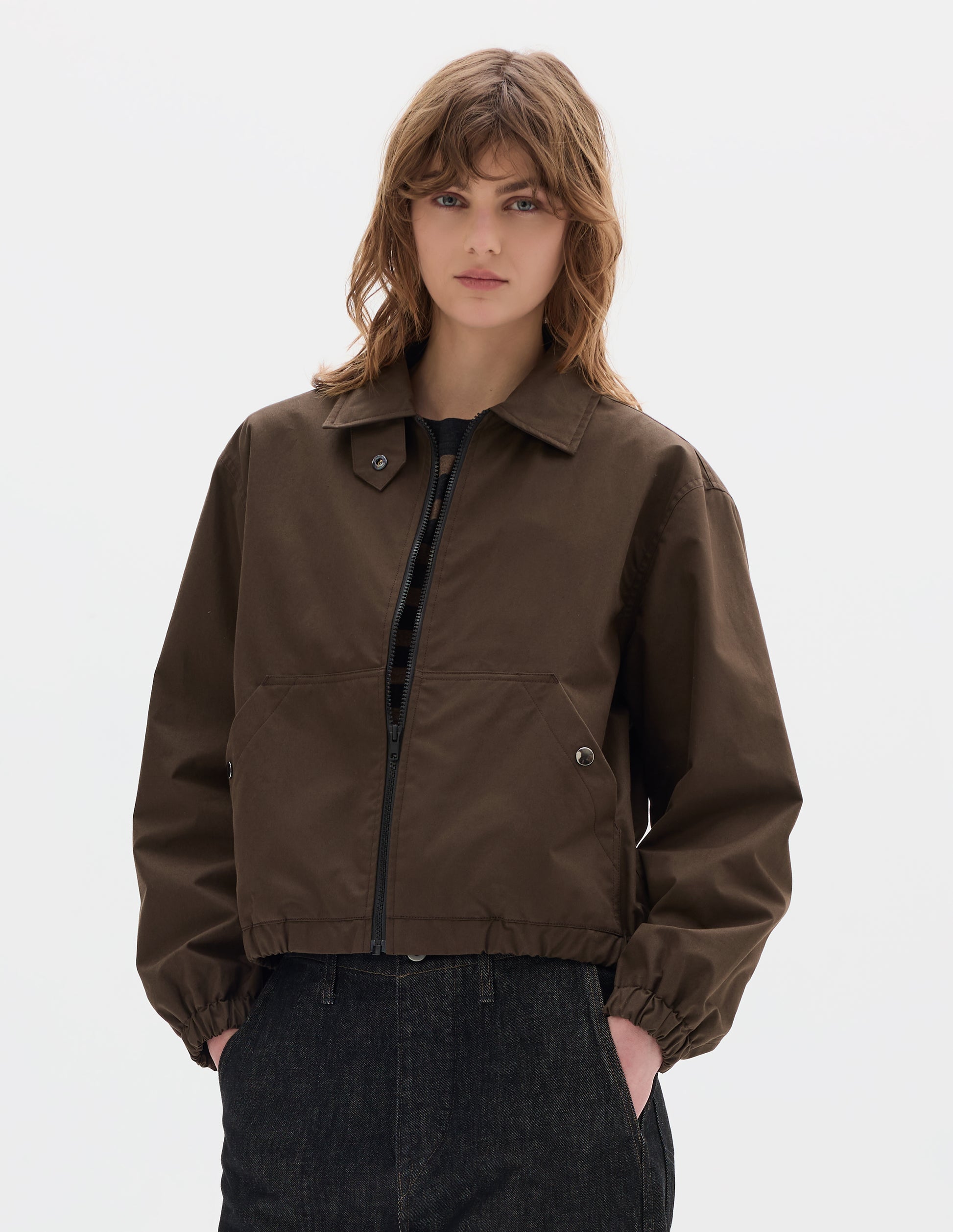 Cropped body image, front view of model wearing an open brown zip-through front jacket, cropped in length, a collar with a throat tab and press stud fastening, two slanted front pockets fastened with press studs, and an elasticated half-hem and cuffs. Styled with black denim.