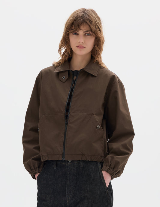 Cropped body image, front view of model wearing an open brown zip-through front jacket, cropped in length, a collar with a throat tab and press stud fastening, two slanted front pockets fastened with press studs, and an elasticated half-hem and cuffs. Styled with black denim.