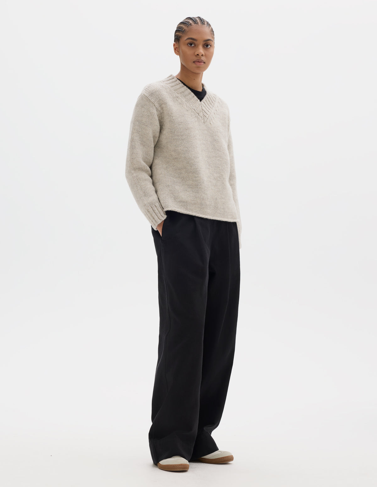 Full body image, front view of model wearing a greyish light beige wool chunky wool sweater with a deep ribbed v neck, relaxed sleeves with rib cuffs and cast-off hem. Styled over a black jersey and with black pleated trousers, off white suede trainers.