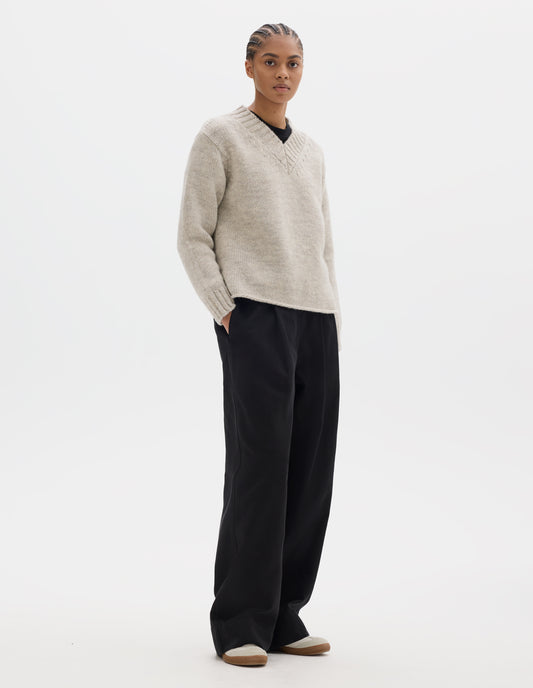 Full body image, front view of model wearing a greyish light beige wool chunky wool sweater with a deep ribbed v neck, relaxed sleeves with rib cuffs and cast-off hem. Styled over a black jersey and with black pleated trousers, off white suede trainers.