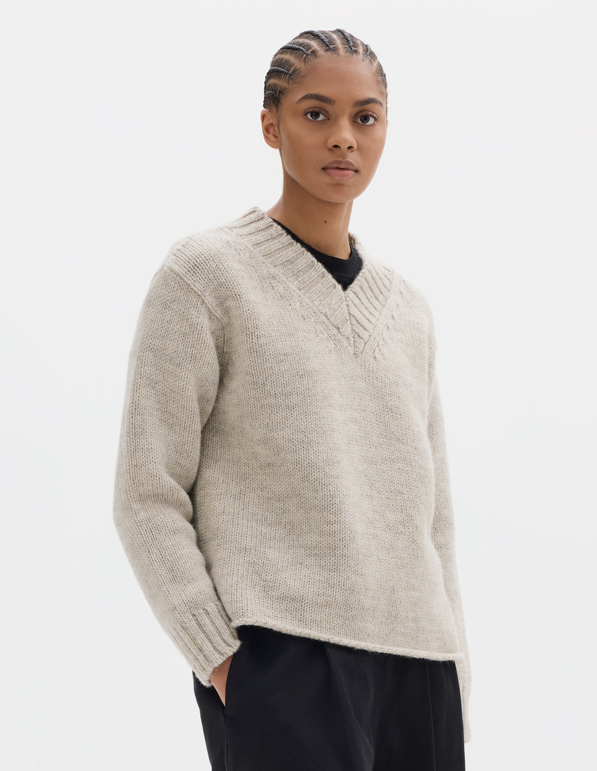 Cropped body image, front view of model wearing a greyish light beige wool chunky wool sweater with a deep ribbed v neck, relaxed sleeves with rib cuffs and cast-off hem. Styled over a black jersey and with black pleated trousers.