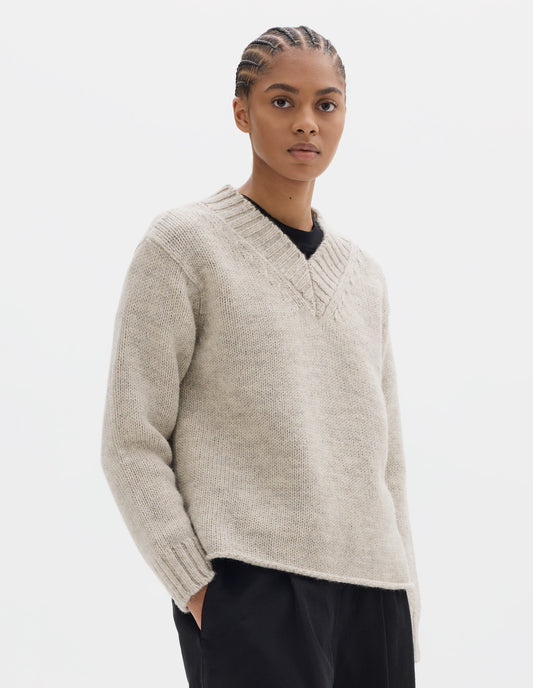 Cropped body image, front view of model wearing a greyish light beige wool chunky wool sweater with a deep ribbed v neck, relaxed sleeves with rib cuffs and cast-off hem. Styled over a black jersey and with black pleated trousers.