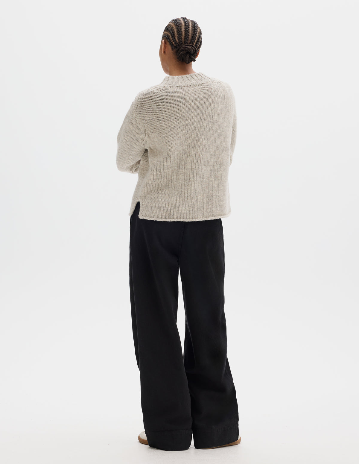Full body image, back view of model wearing a greyish light beige wool chunky wool sweater with a deep ribbed neck, relaxed sleeves, cast-off hem with side splits. Styled with black trousers, off white suede trainers.