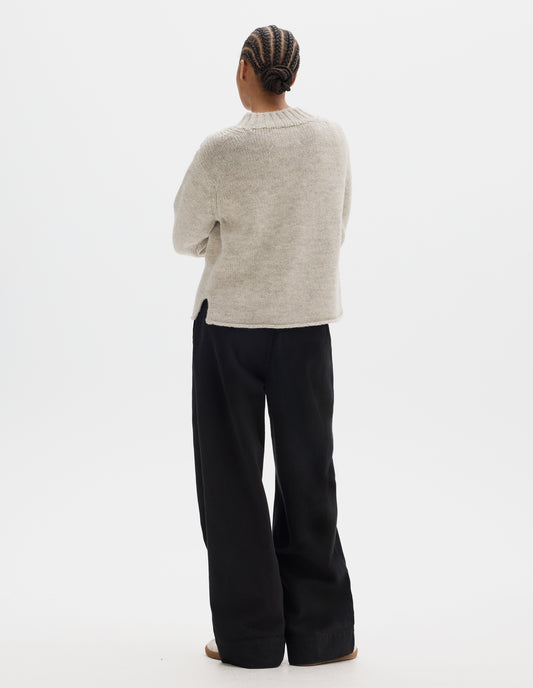 Full body image, back view of model wearing a greyish light beige wool chunky wool sweater with a deep ribbed neck, relaxed sleeves, cast-off hem with side splits. Styled with black trousers, off white suede trainers.
