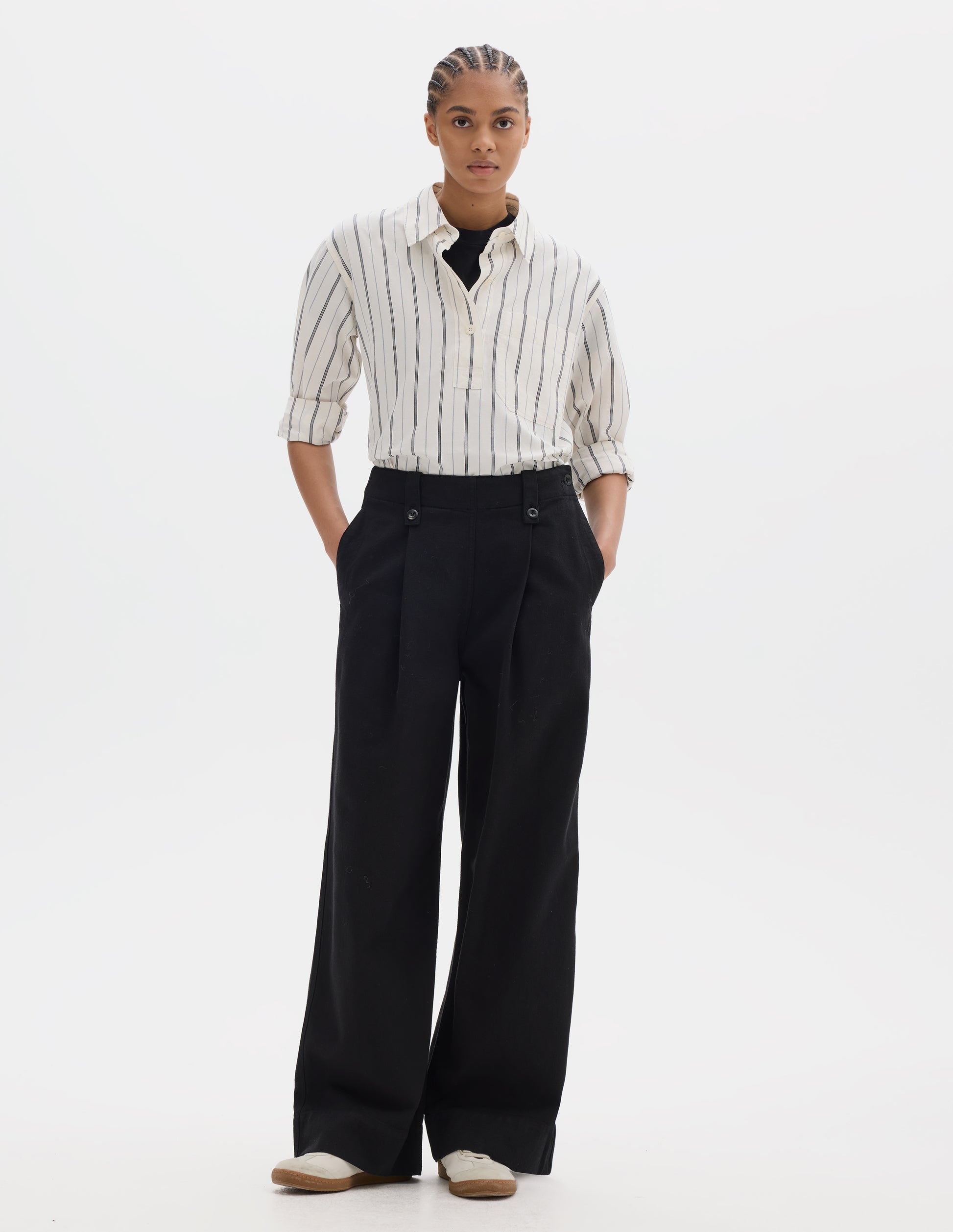 Full body image, front view of model wearing a black wide leg trousers with a button-fastening belt loops, a side opening with integrated side seam pockets and two front pleats. Styled with an off white and dark blue stripe cotton shirt with light buttons, a half placket, worn over a black t shirt and off white suede trainers.