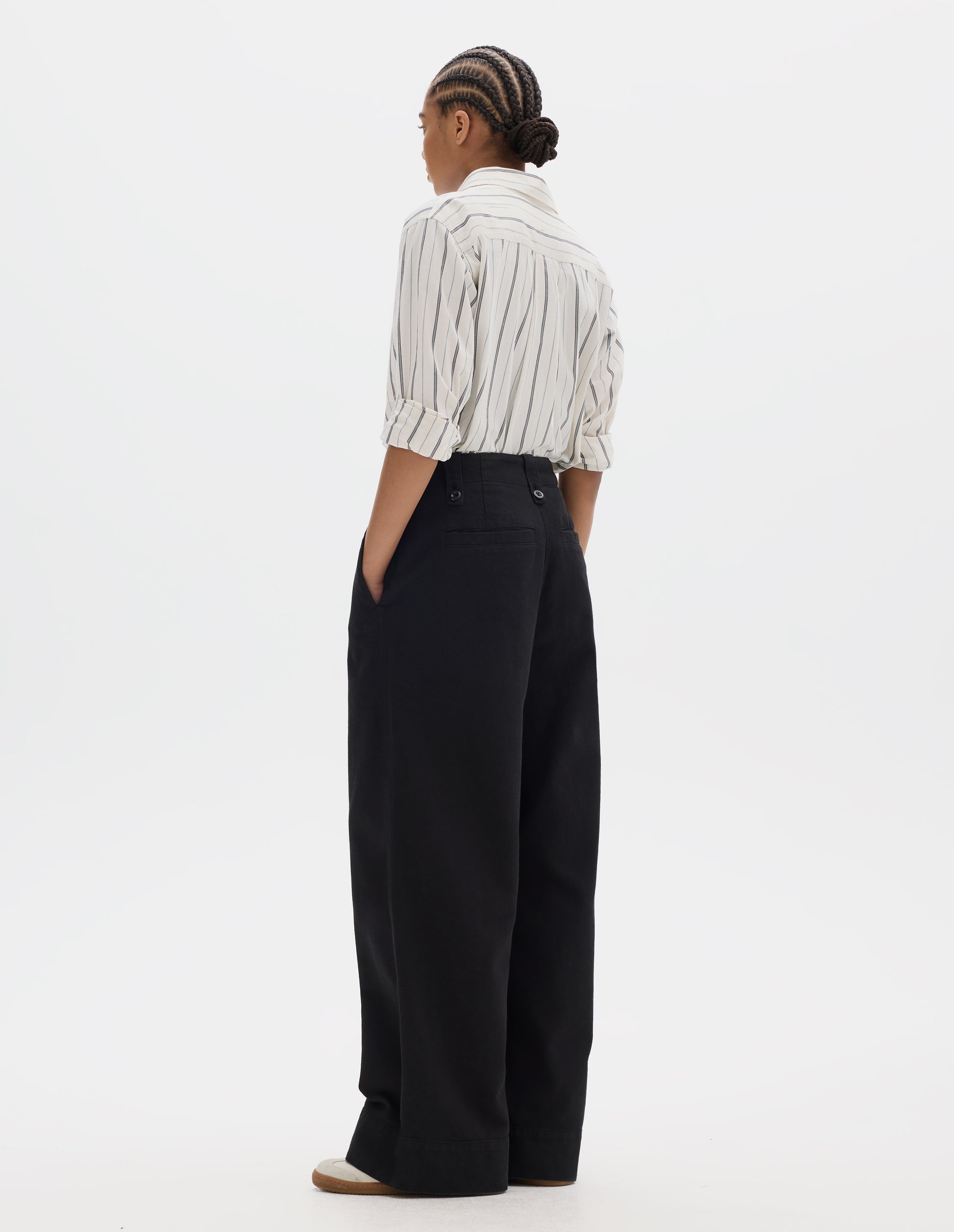 Full body image, back view of model wearing a black wide leg trousers with a button-fastening belt loops, a side opening with integrated side seam pockets and back welt pockets and a deep stitch-through hem. Styled with an off white and dark blue stripe cotton shirt and off white suede trainers.