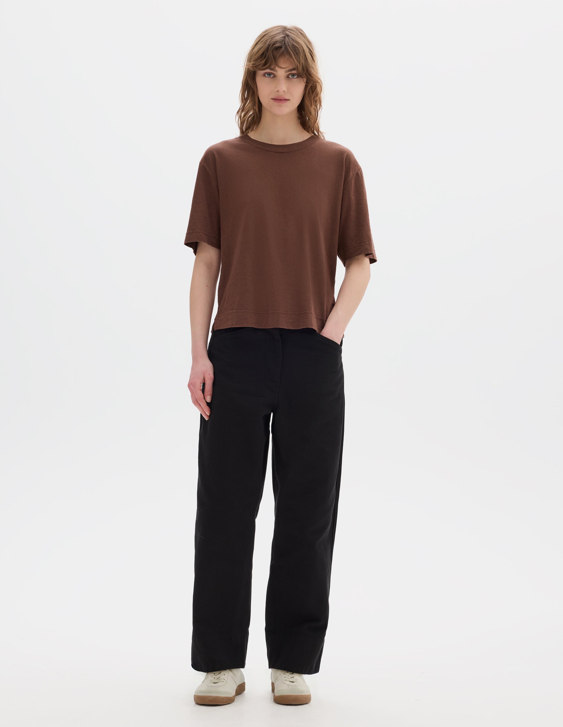 Full body image, front view of model wearing brown cotton t shirt, with side vents. Styled with black trousers and white shoes. 