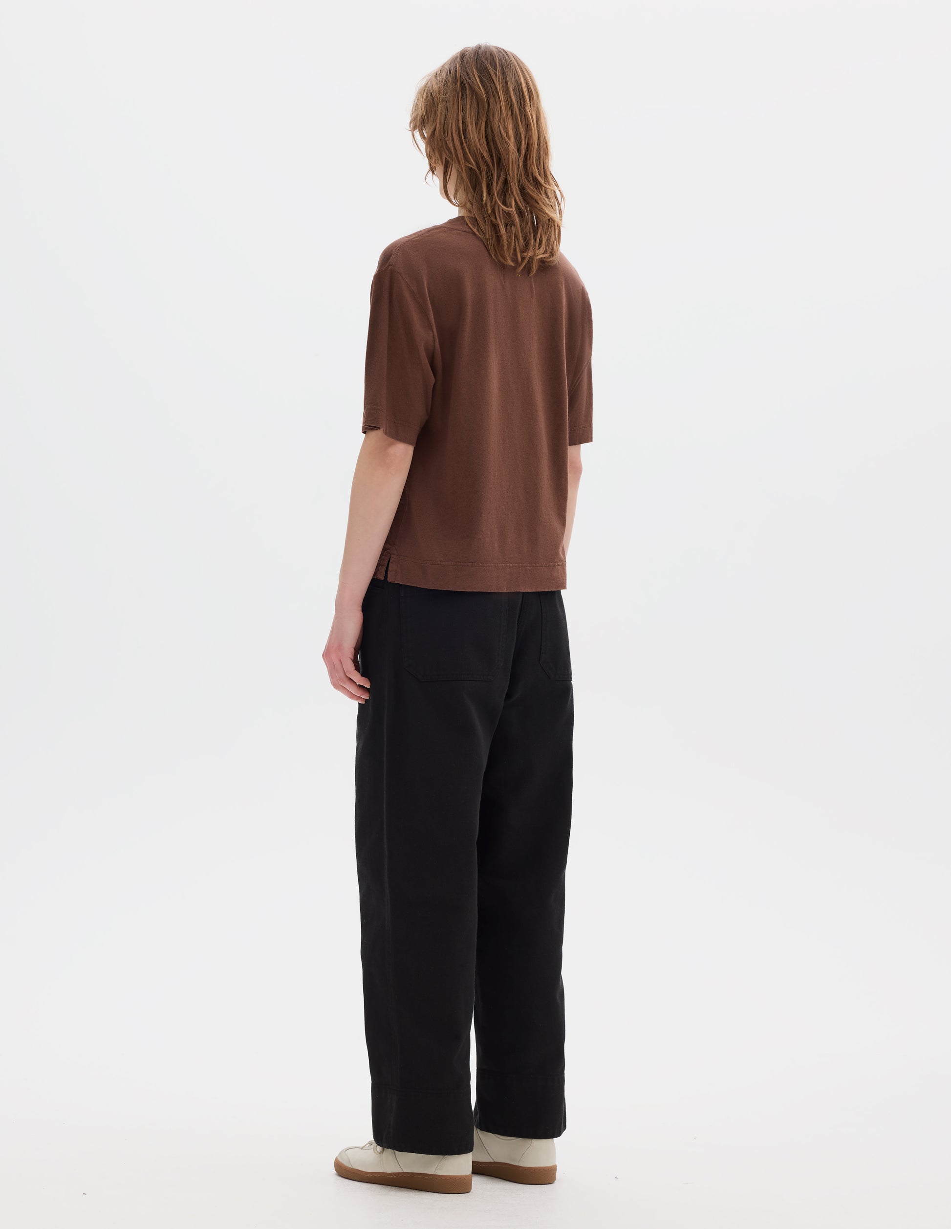 Full body image, back view of model wearing brown cotton t shirt, with side vents. Styled with black trousers and white shoes. 