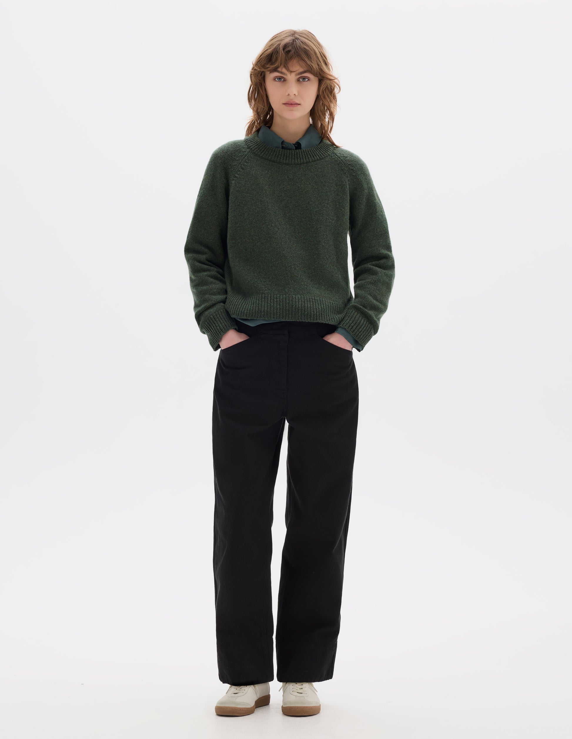 Full body image, front view of model wearing green knitted wool jumper with crew neck, raglan sleeves, and rib detailing on the neck, cuffs and hem. Styled over green cotton shirt, with black cotton trousers and white trainers with gum sole.