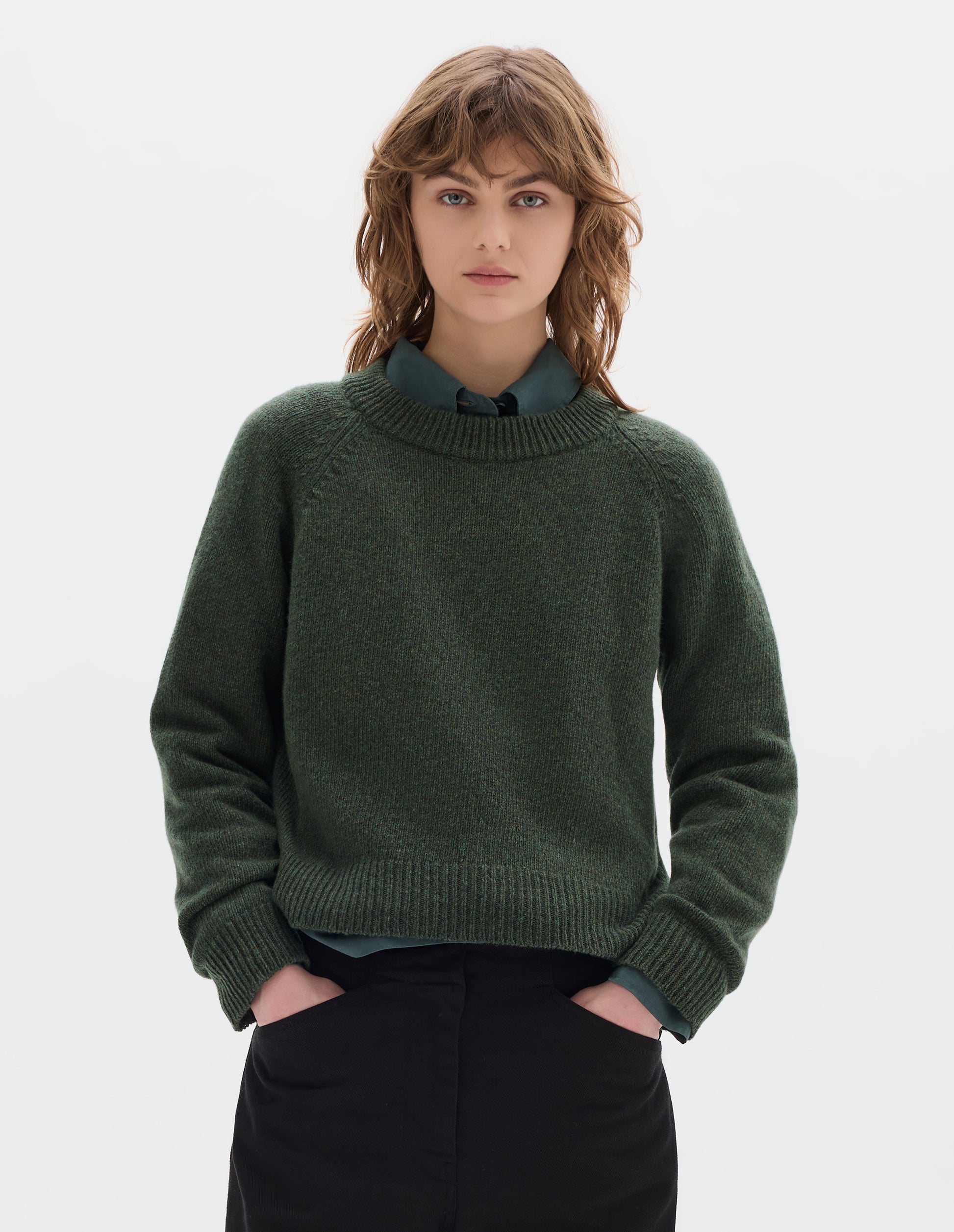 Cropped body image, front view of model wearing green knitted wool jumper with crew neck, raglan sleeves, and rib detailing on the neck, cuffs and hem. Styled over green cotton shirt, with black cotton trousers.