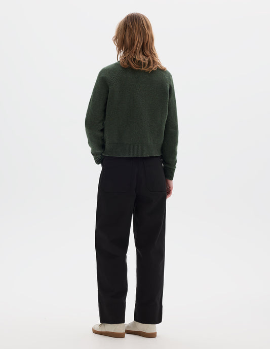 Full body image, back view of model wearing green knitted wool jumper with raglan sleeves, and rib detailing on the neck, cuffs and hem. Styled with black cotton trousers and white trainers with gum sole.